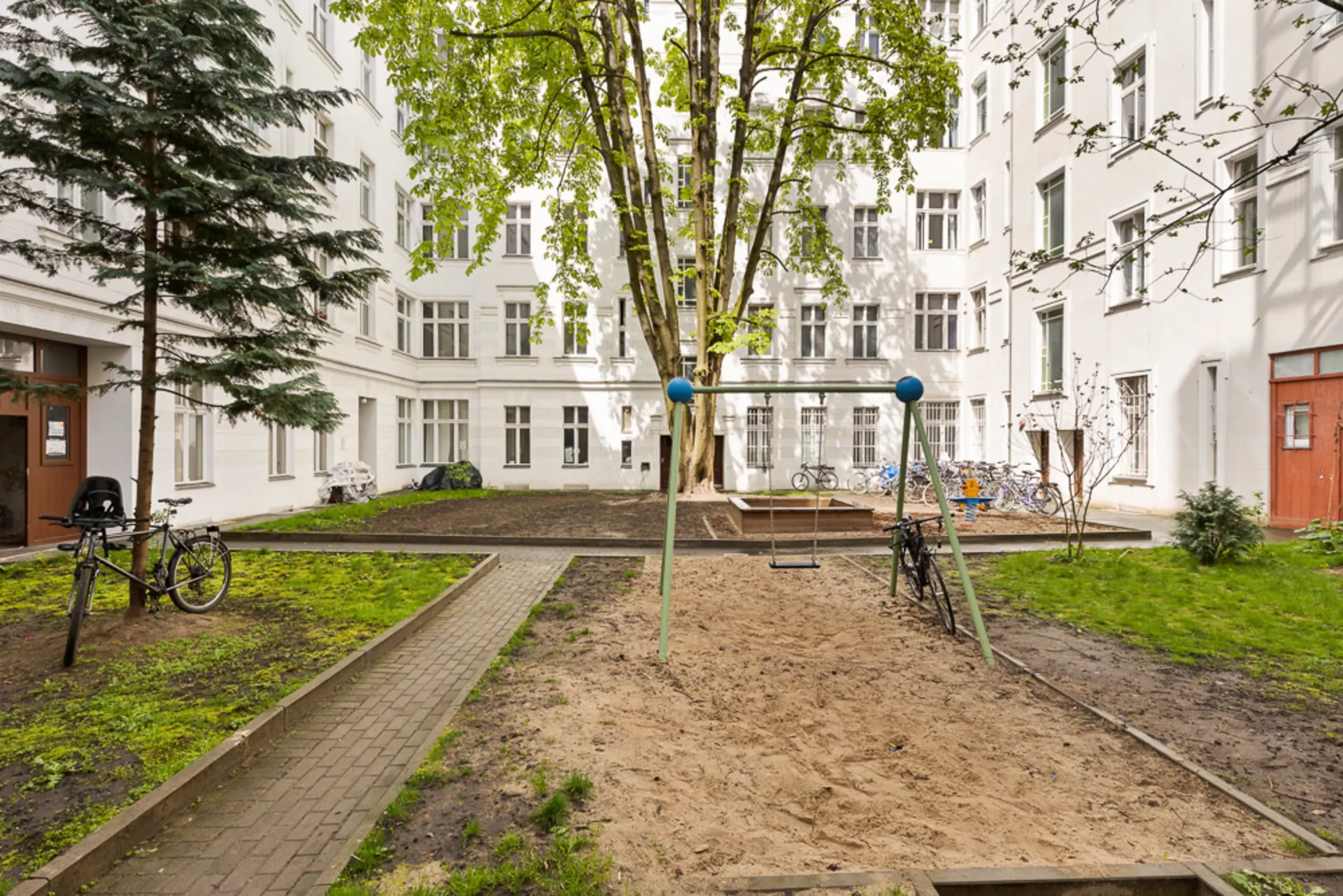 Innenhof mit Schaukel, Sandfläche, Fahrrädern und Bäumen, umgeben von weißen Wohngebäuden.