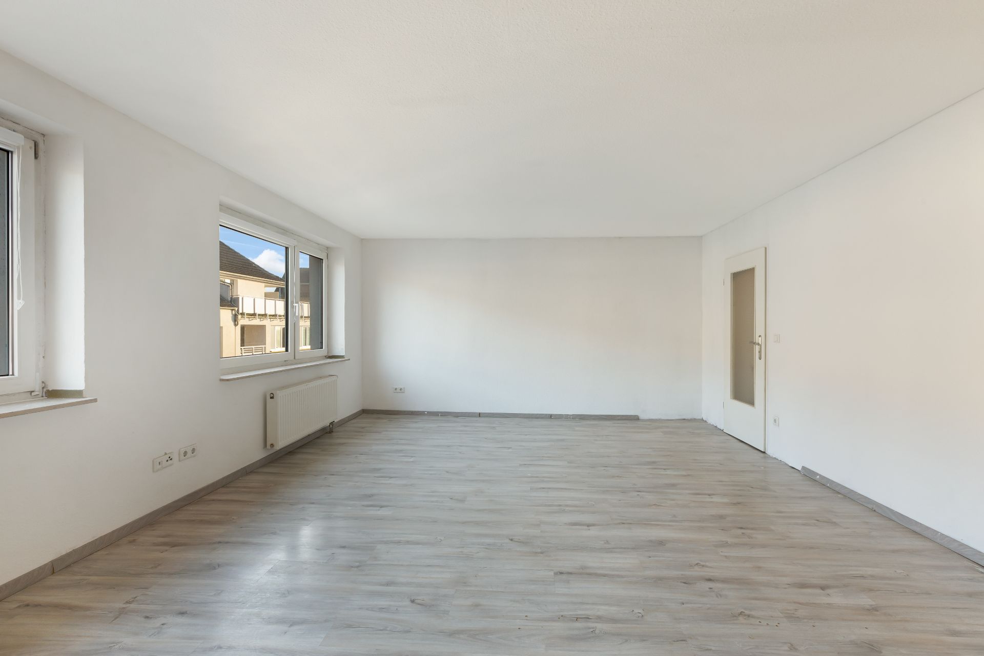Heller, leerer Wohnraum mit zwei Fenstern, weiß gestrichenen Wänden und Laminatboden.