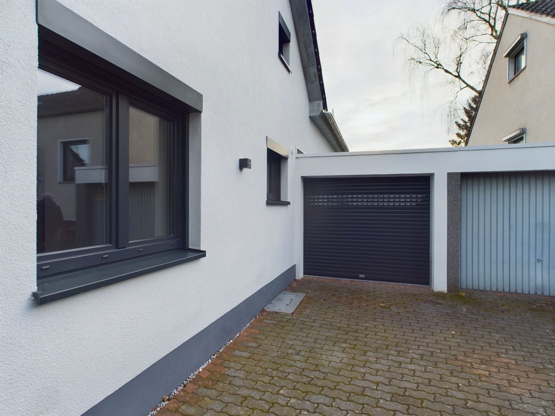 Gepflasterter Hof mit weißer Hauswand, großem Fenster und zwei Garagentoren.
