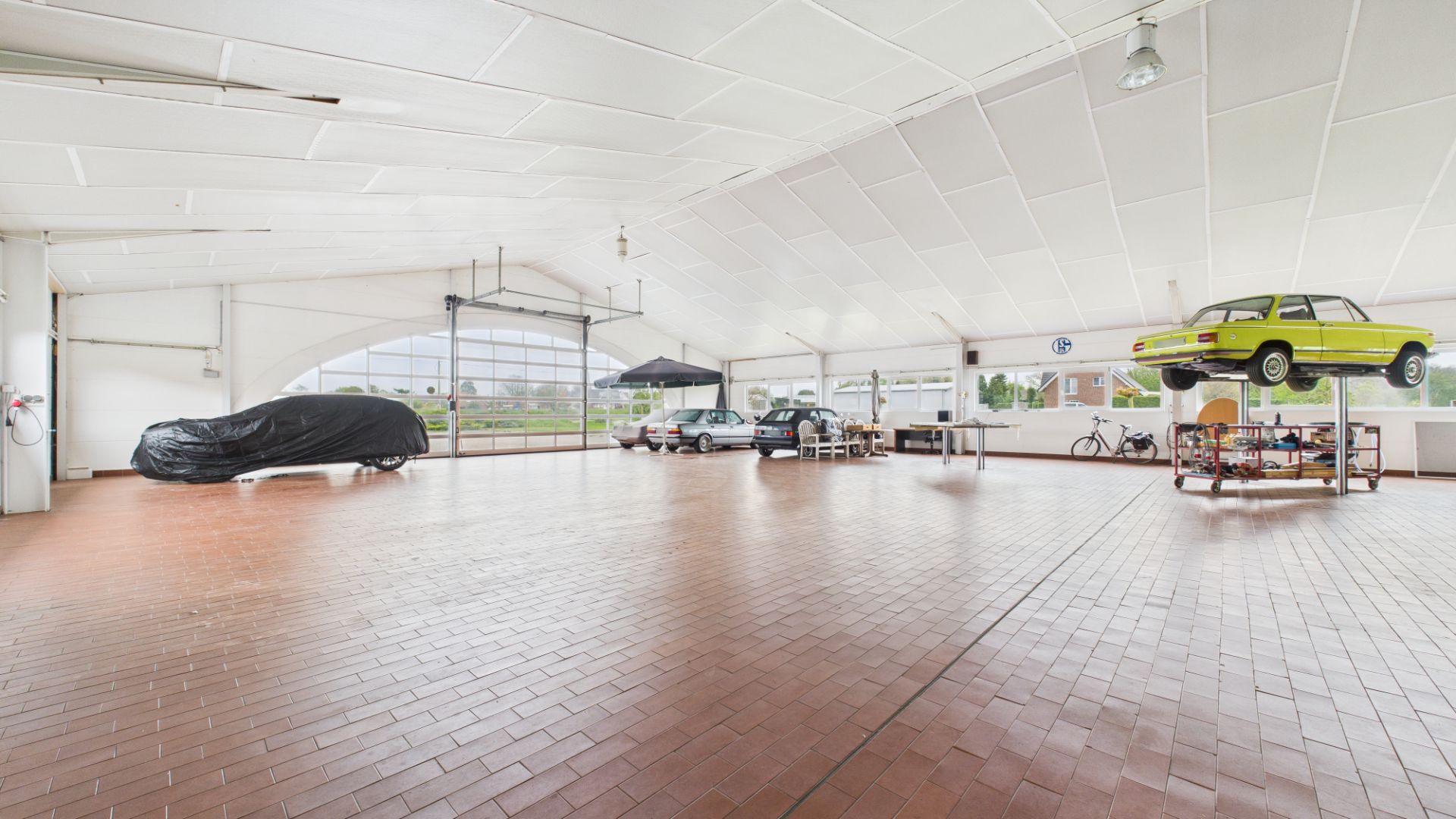 Große helle Garage mit Autos, darunter ein angehobenes gelbes Auto und ein Auto unter einer Abdeckung.