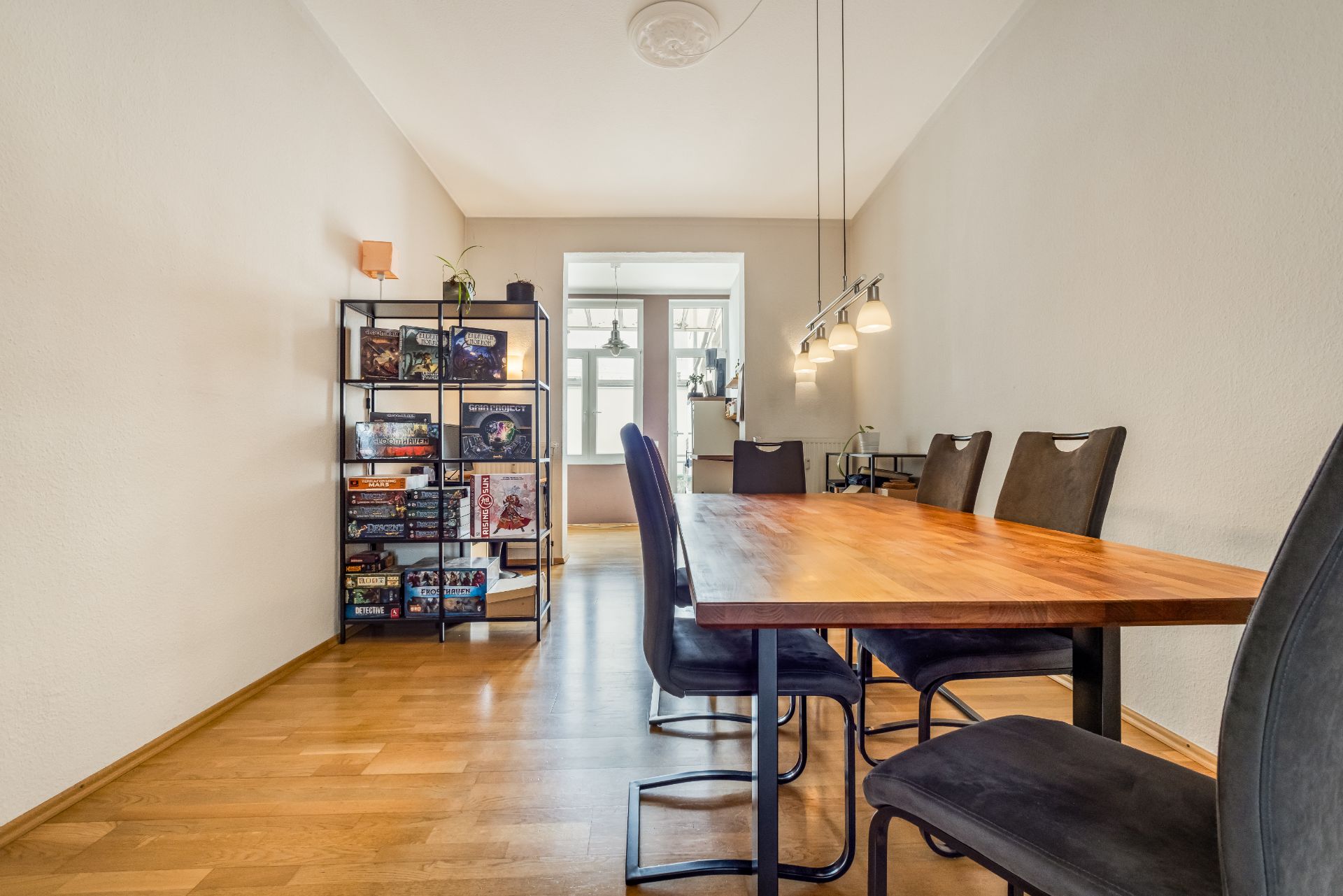 Esszimmer mit Holztisch, sechs Stühlen, Bücherregal und moderner Hängelampe auf Parkettboden.