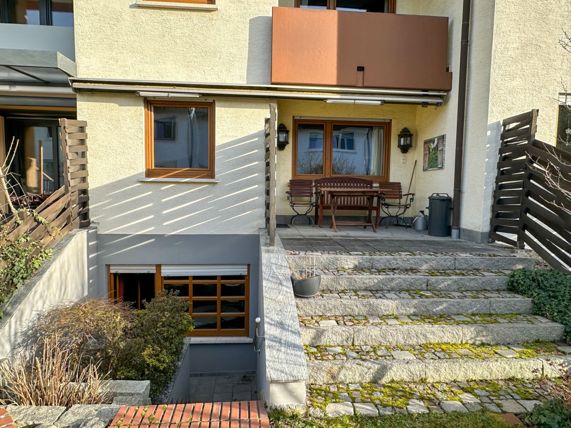 Steintreppe führt zu einer Terrasse mit Holzmöbeln vor einem Wohnhaus, darunter ein Kellereingang.