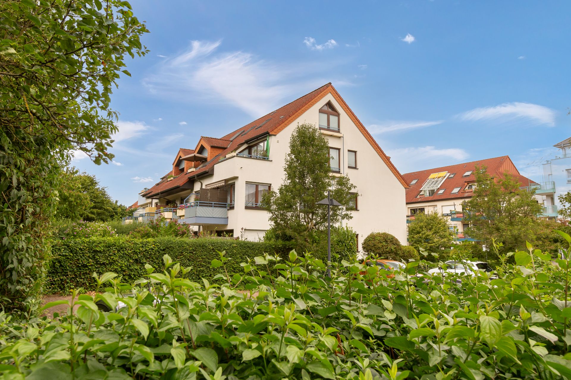 Mehrfamilienhaus mit Balkonen und Satteldach, umgeben von Büschen und Bäumen.