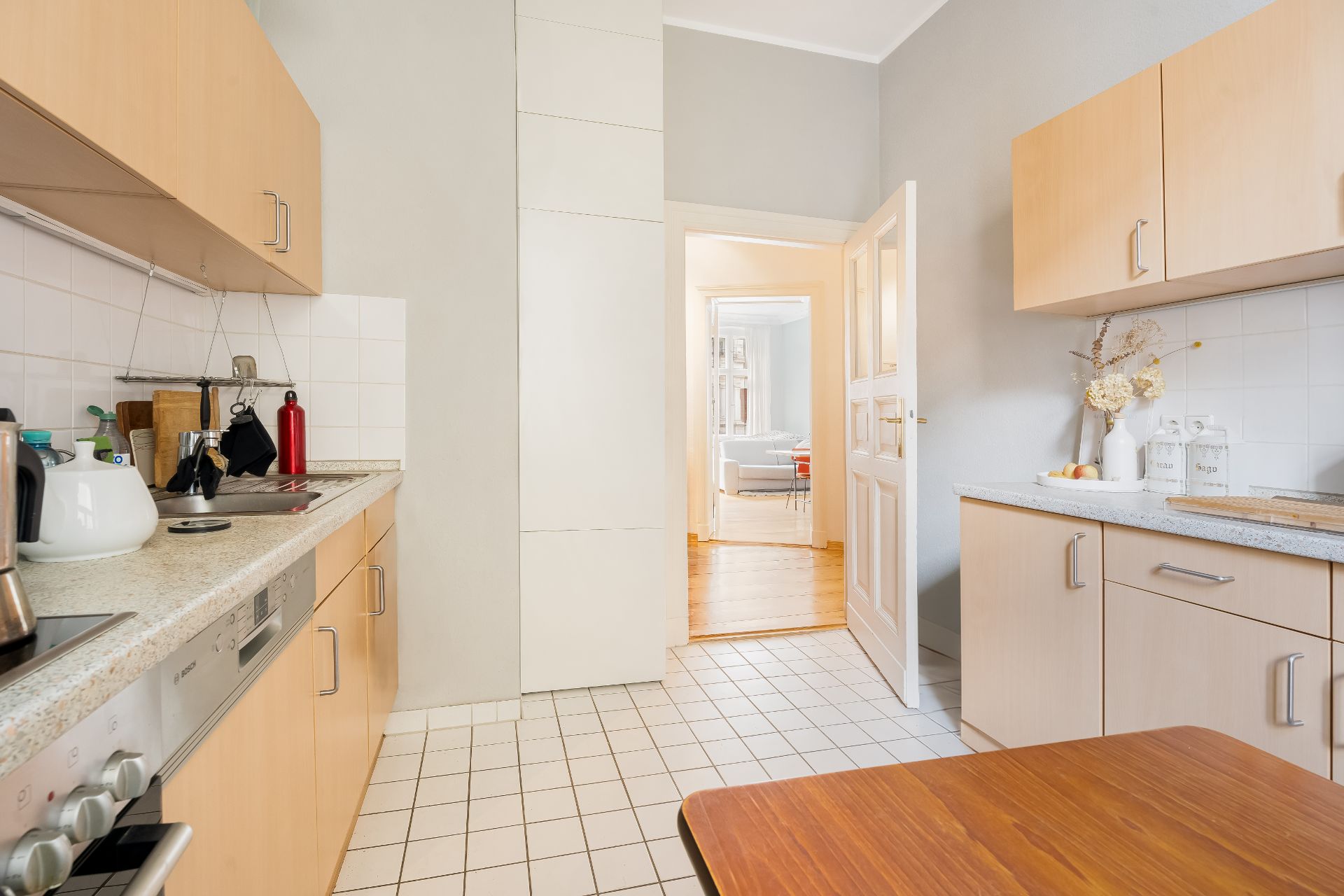 Helle Küche mit Holzmöbeln, Fliesenboden und Blick in ein Wohnzimmer mit Sofa.