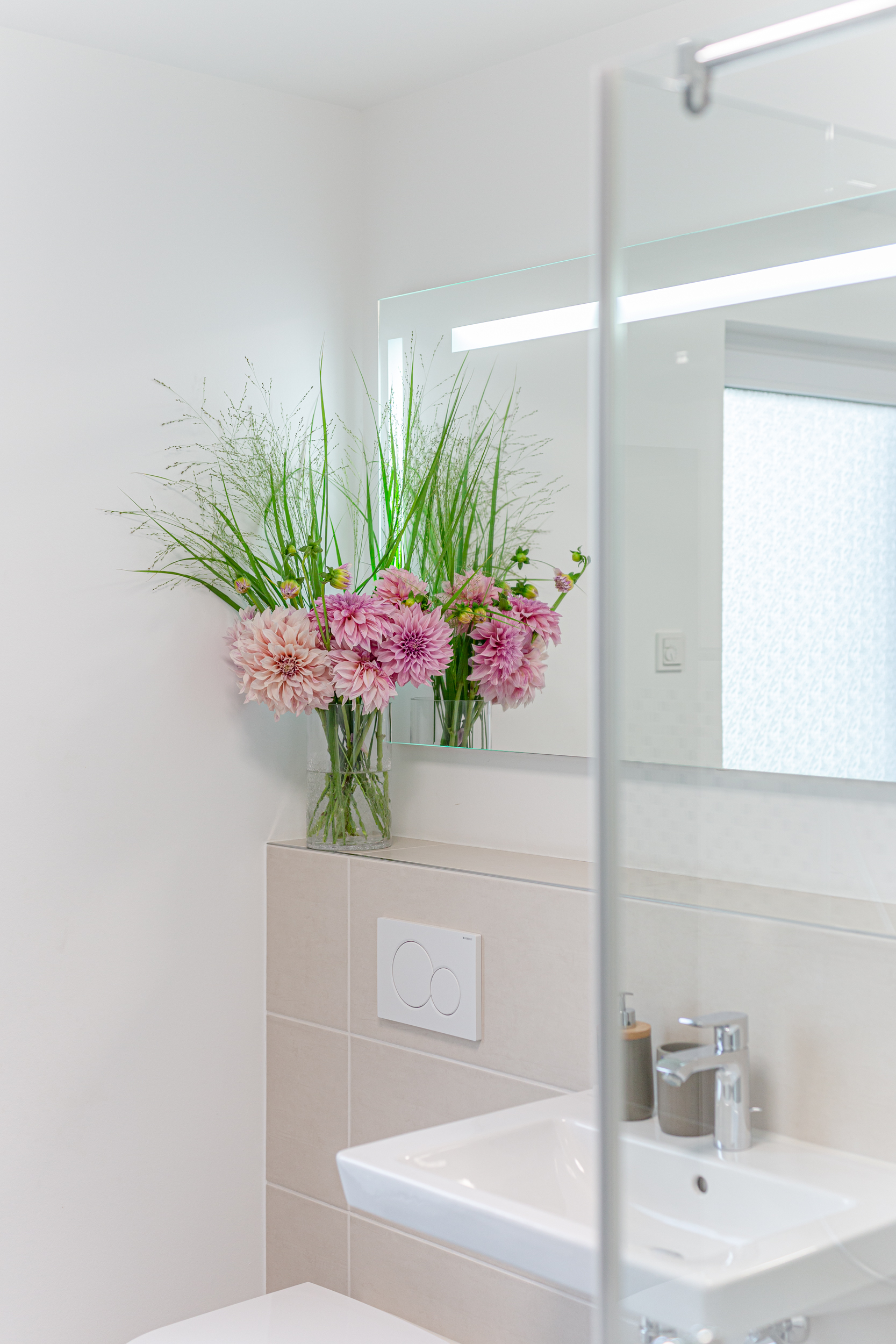 Modernes Bad mit weißem Waschbecken, Spiegel und Vase mit rosa Blumen auf der Ablage.