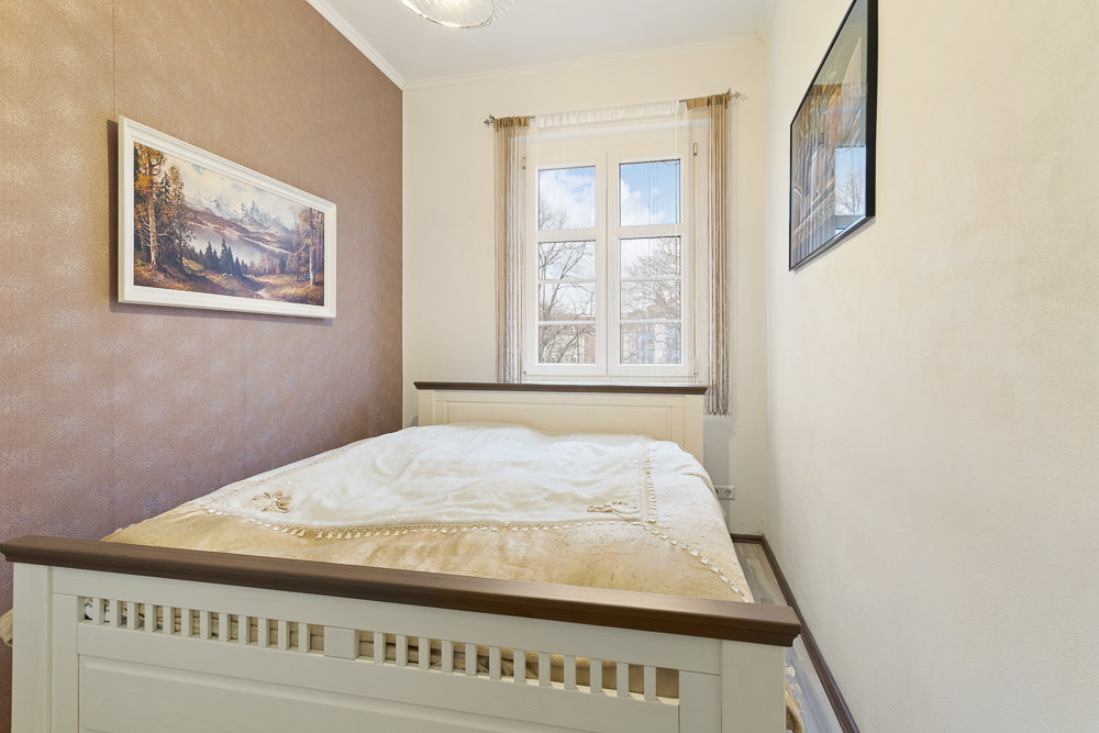 Helles Schlafzimmer mit Bett, Fenster, Gemälde an den Wänden und heller Bettdecke.