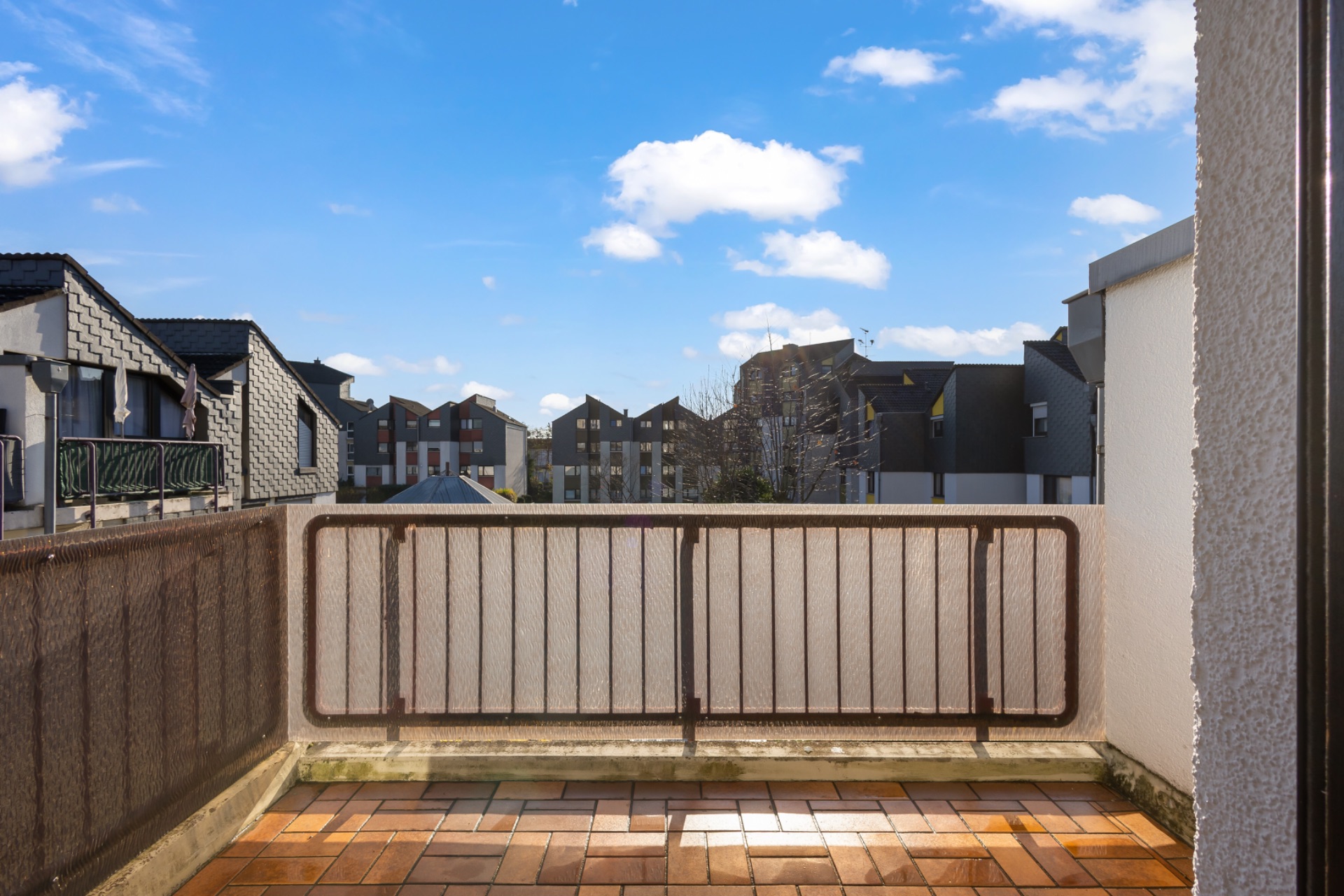 Balkon mit braunem Geländer und Fliesenboden, Blick auf angrenzende Wohnhäuser unter blauem Himmel.