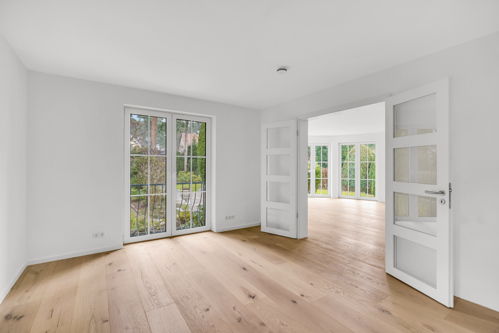 Helles Zimmer mit Holzboden, Fenster und Doppeltüren zu einem weiteren Raum mit großen Fenstern.