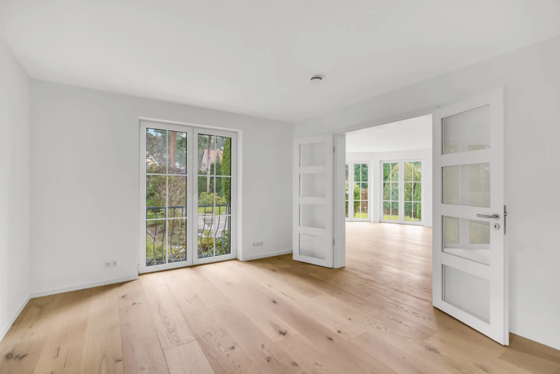 Helles Zimmer mit Holzboden, Fenster und Doppeltüren zu einem weiteren Raum mit großen Fenstern.