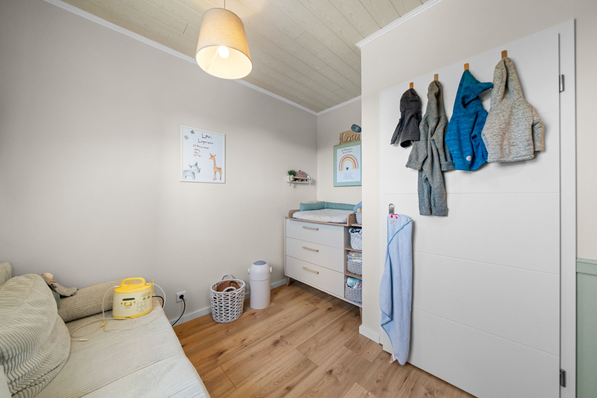 Helles Kinderzimmer mit Wickelkommode, Sofa, Kleiderhaken und Bildern an der Wand.