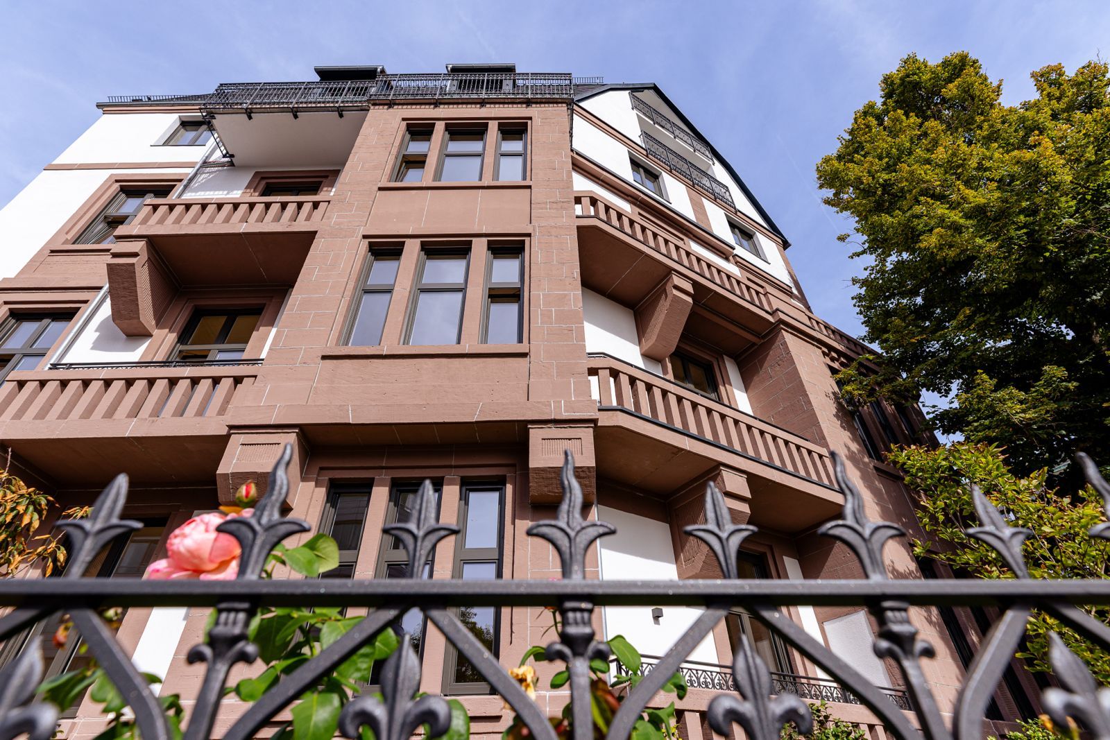 Mehrstöckiges Wohnhaus mit braunen Fenstern und Balkonen, Blick über einen Zaun und Blumen.