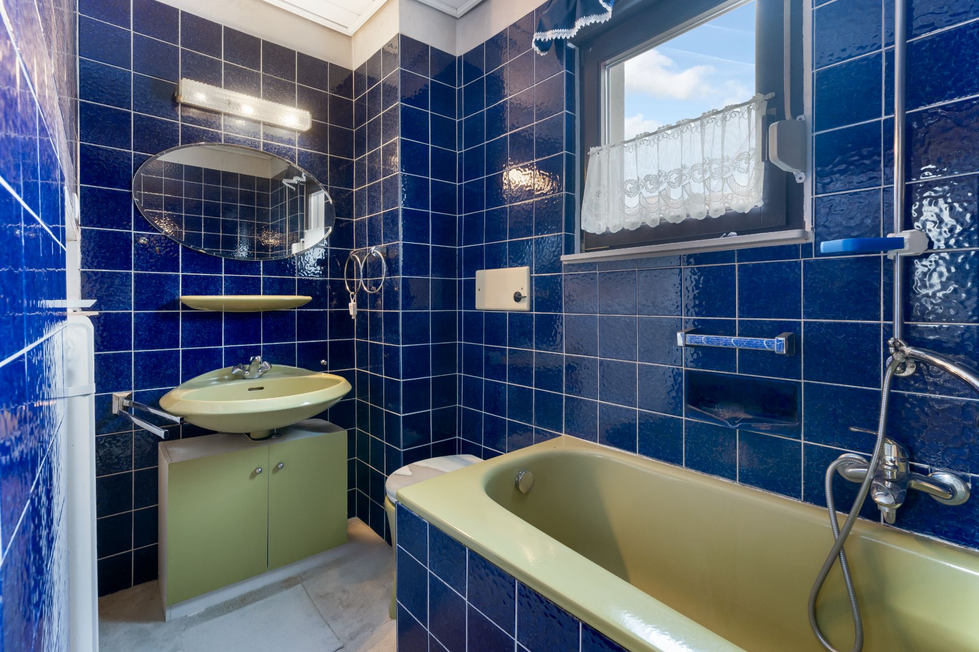 Kleines Bad mit blauen Fliesen, grünem Waschbecken und Badewanne, einem Fenster und Wandspiegel.