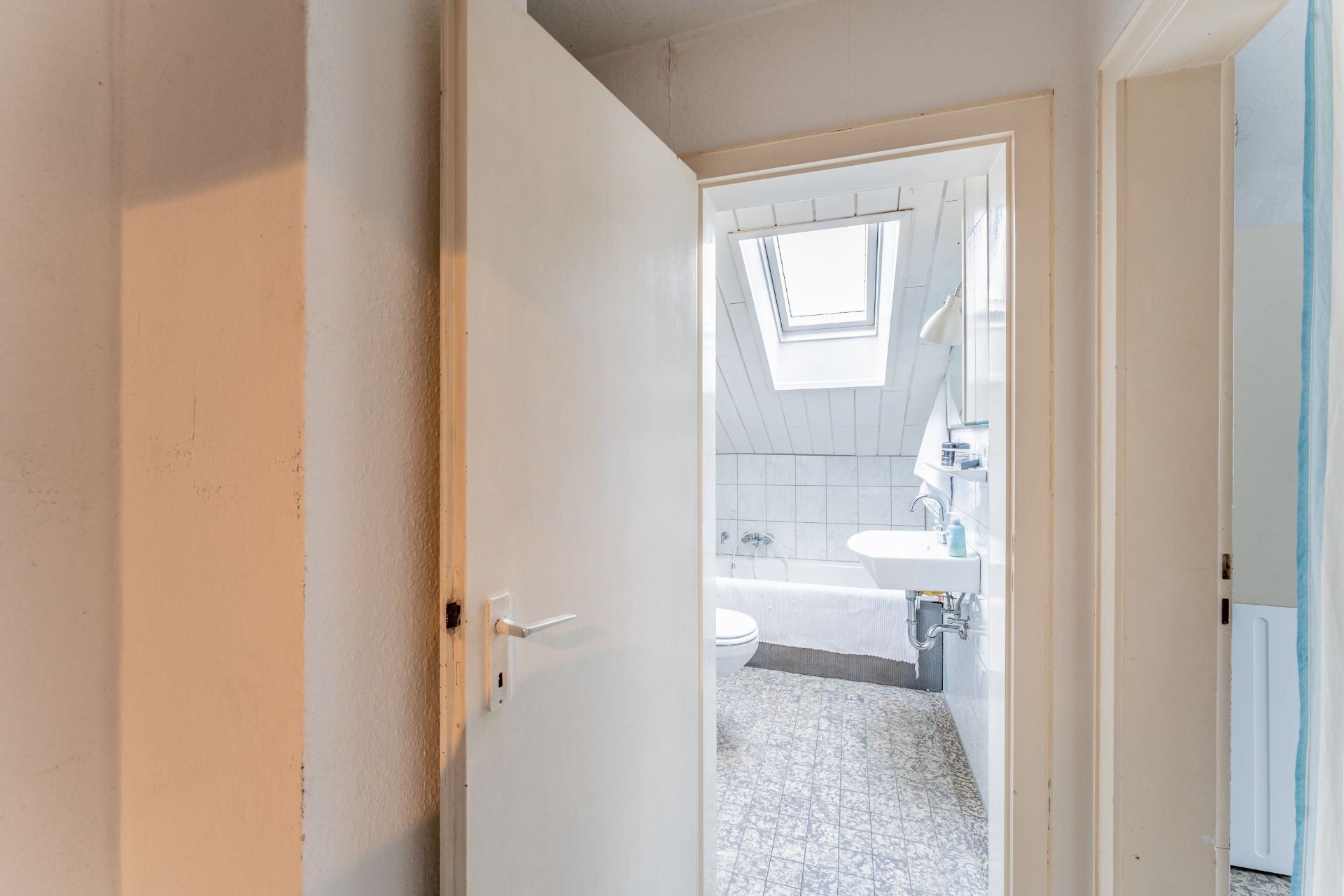 Blick durch offene Tür in ein helles Badezimmer mit Dachfenster, Waschbecken und Badewanne.