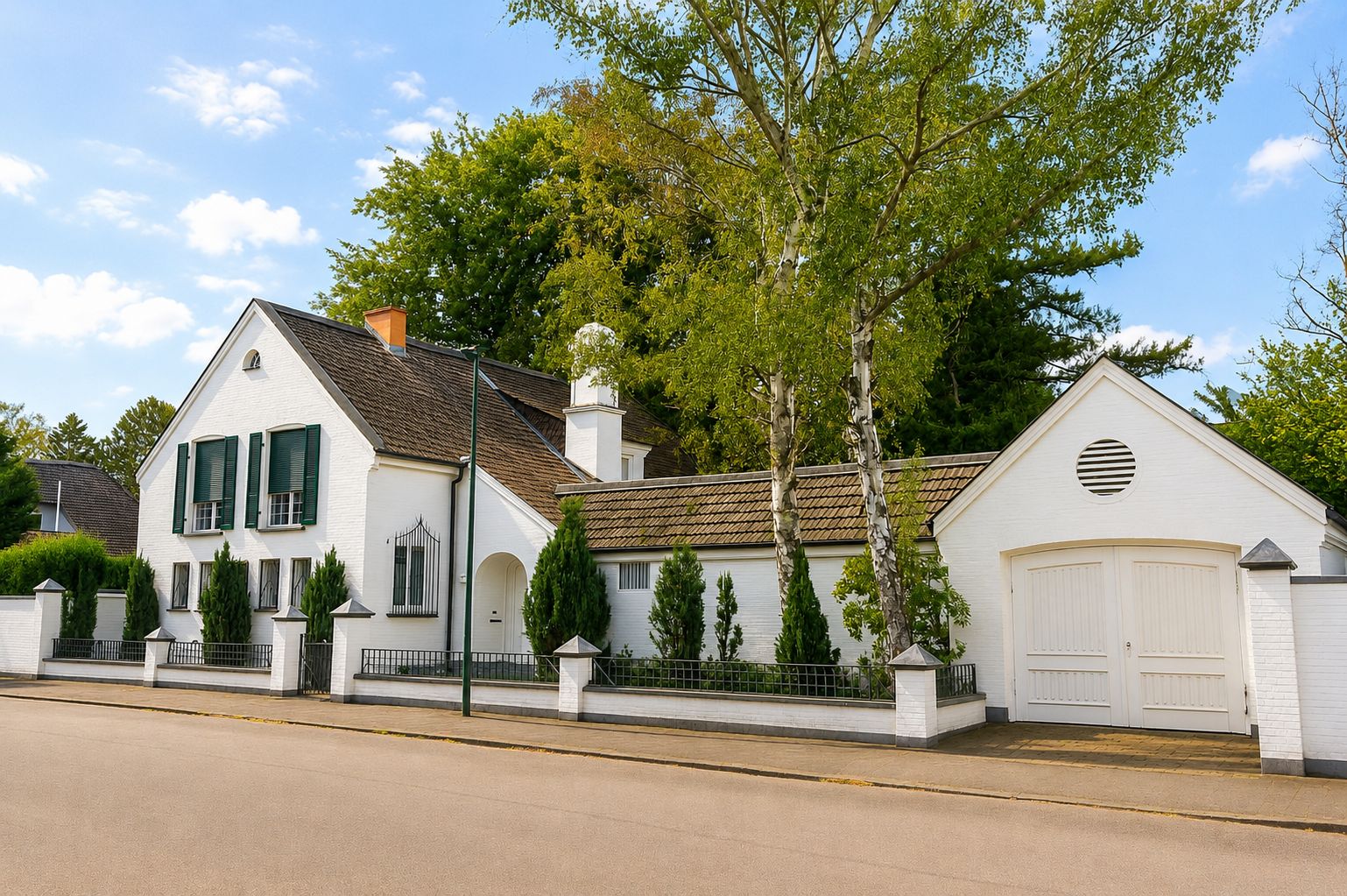 Weißes Wohnhaus mit grünem Fensterläden, Garage und Garten vor Bäumen an einer Straße.