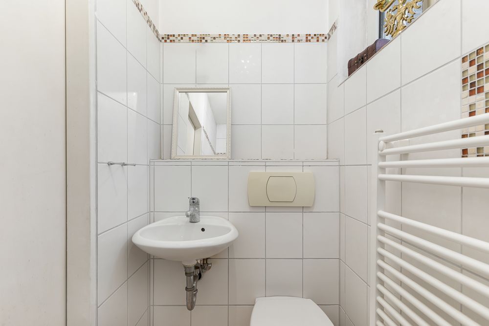 Kleines, weiß gefliestes Bad mit Waschbecken, Spiegel, Toilette und Heizkörper.