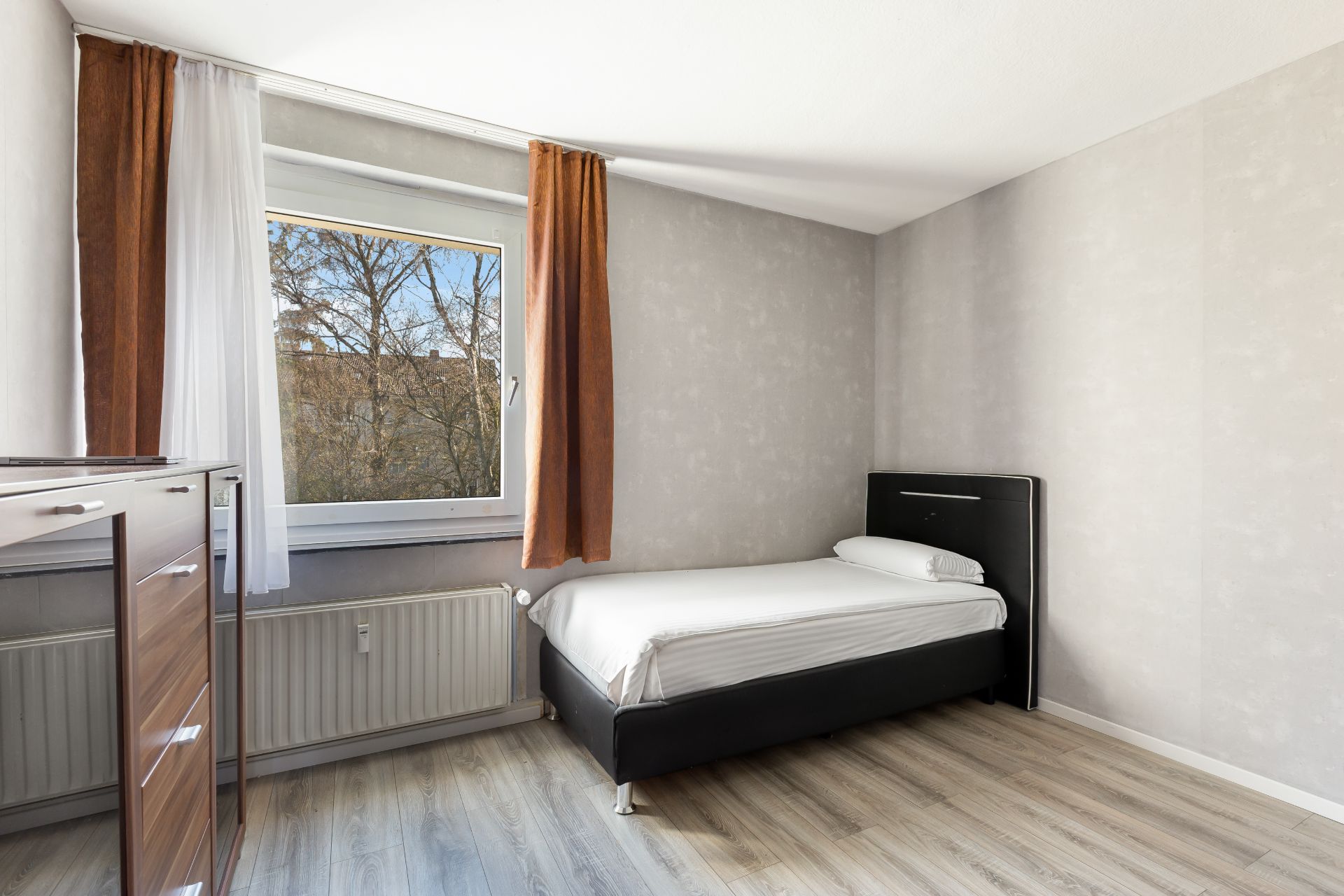 Helles Schlafzimmer mit Einzelbett, Fenster, braunen Vorhängen und Kommode.
