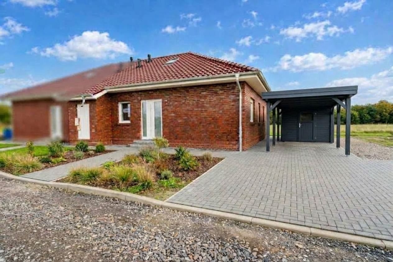 Backsteinhaus mit rotem Dach, gepflasterter Hof und überdachtem Carport, daneben Gartenbeet.