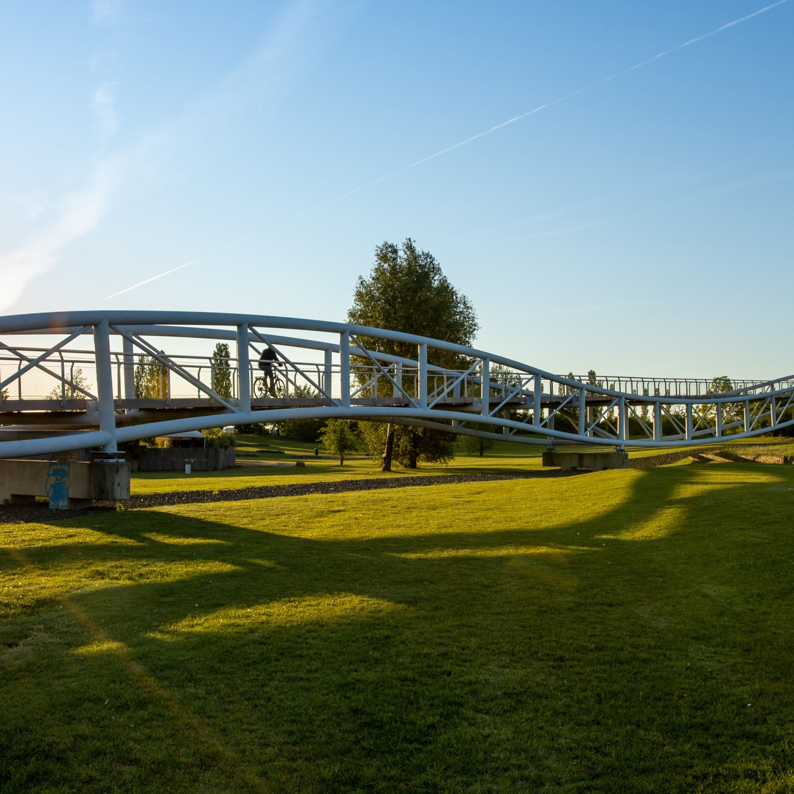 Metallbrücke über einer grünen Wiese bei Sonnenlicht, ein Fahrradfahrer auf der Brücke.