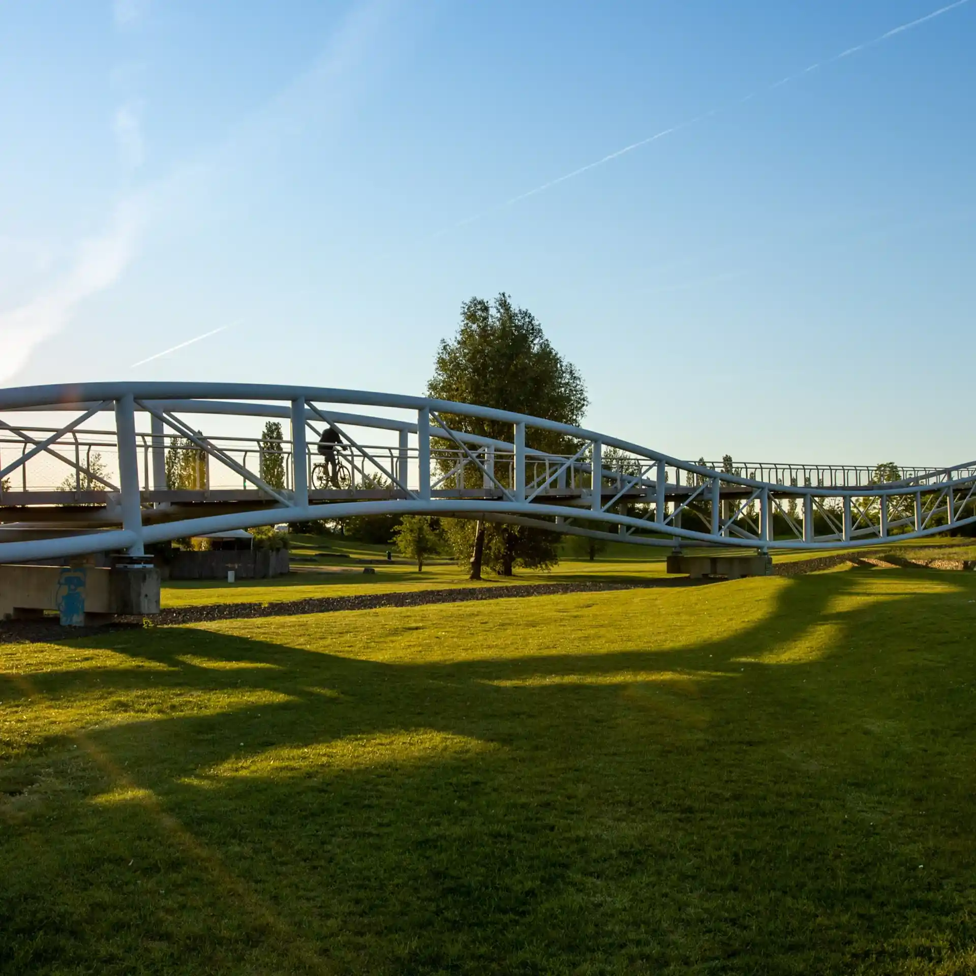 Metallbrücke über einer grünen Wiese bei Sonnenlicht, ein Fahrradfahrer auf der Brücke.
