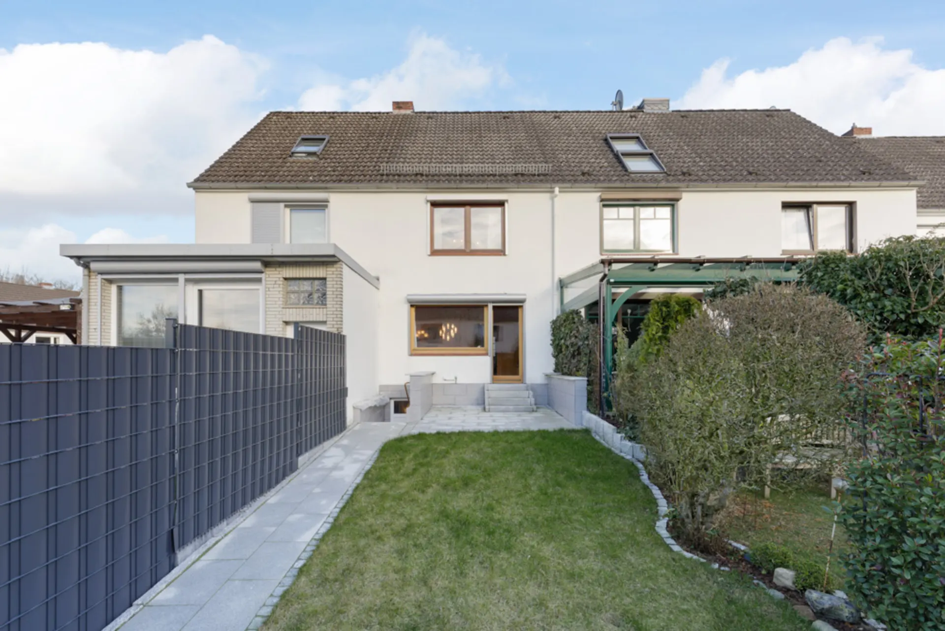 Rückseite eines Reihenhauses mit Garten, Terrasse und Wegen, umgeben von Zäunen und Büschen.