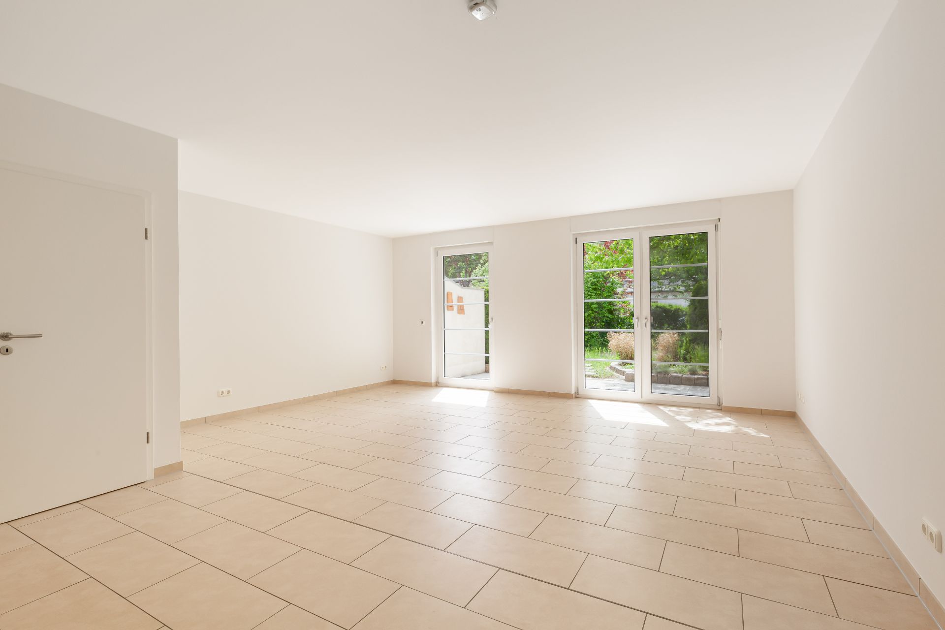 Helles, leeres Zimmer mit Fliesenboden und großen Fenstern zum Garten.