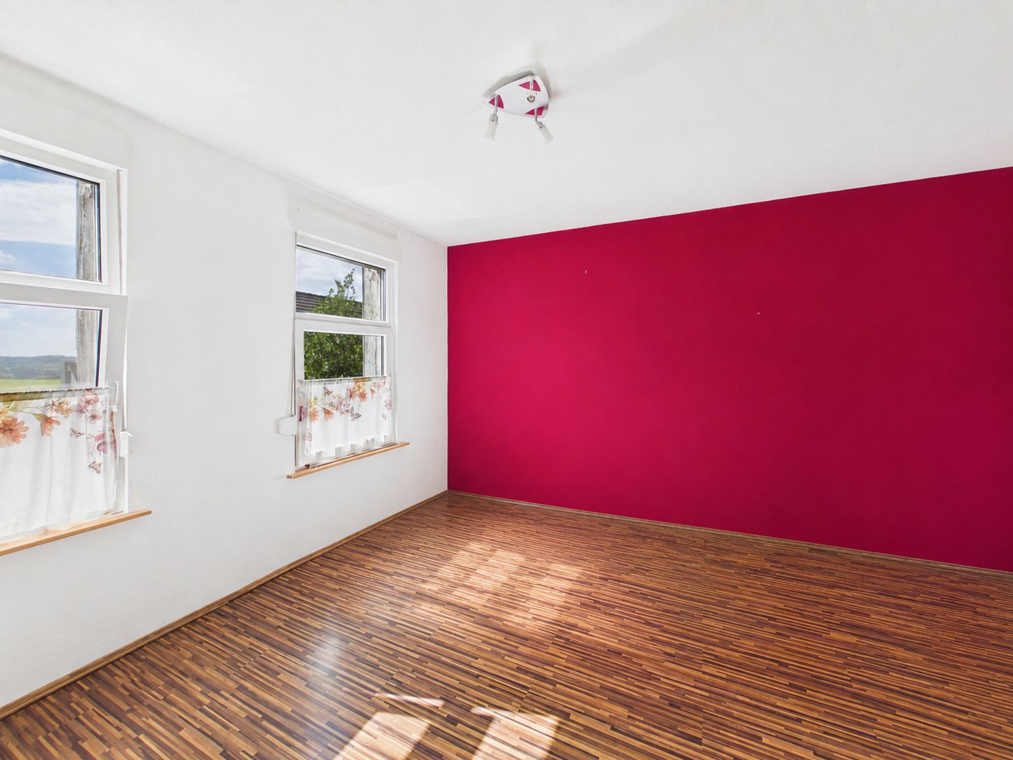 Leerer Raum mit Holzfußboden, zwei Fenstern und einer roten Wand.