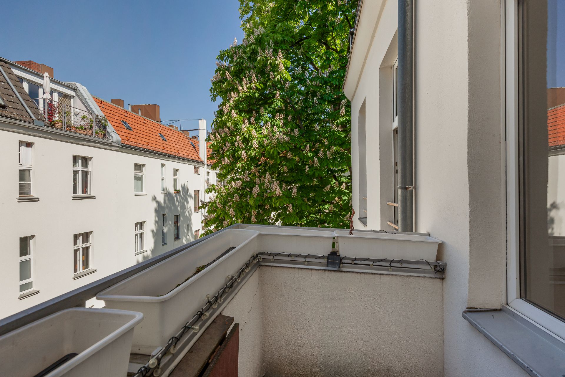 Kleiner Balkon mit weißen Blumenkästen, Ausblick auf Wohnhäuser und grünem Baum.