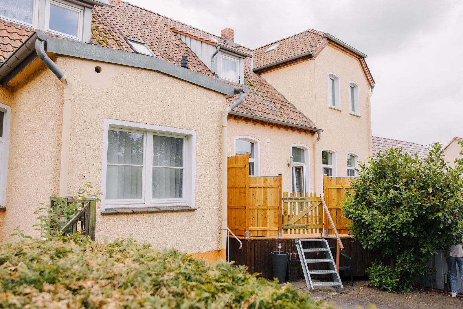 Rückseite eines hellen Hauses mit Terrasse, Holztreppe und kleinen Fenstern im Dach.