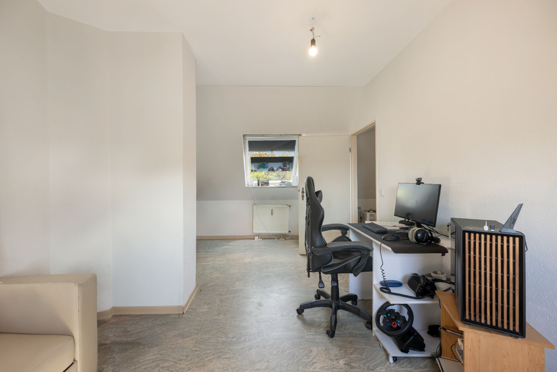 Helles Zimmer mit Schreibtisch, Bürostuhl, Computer und Fenster.