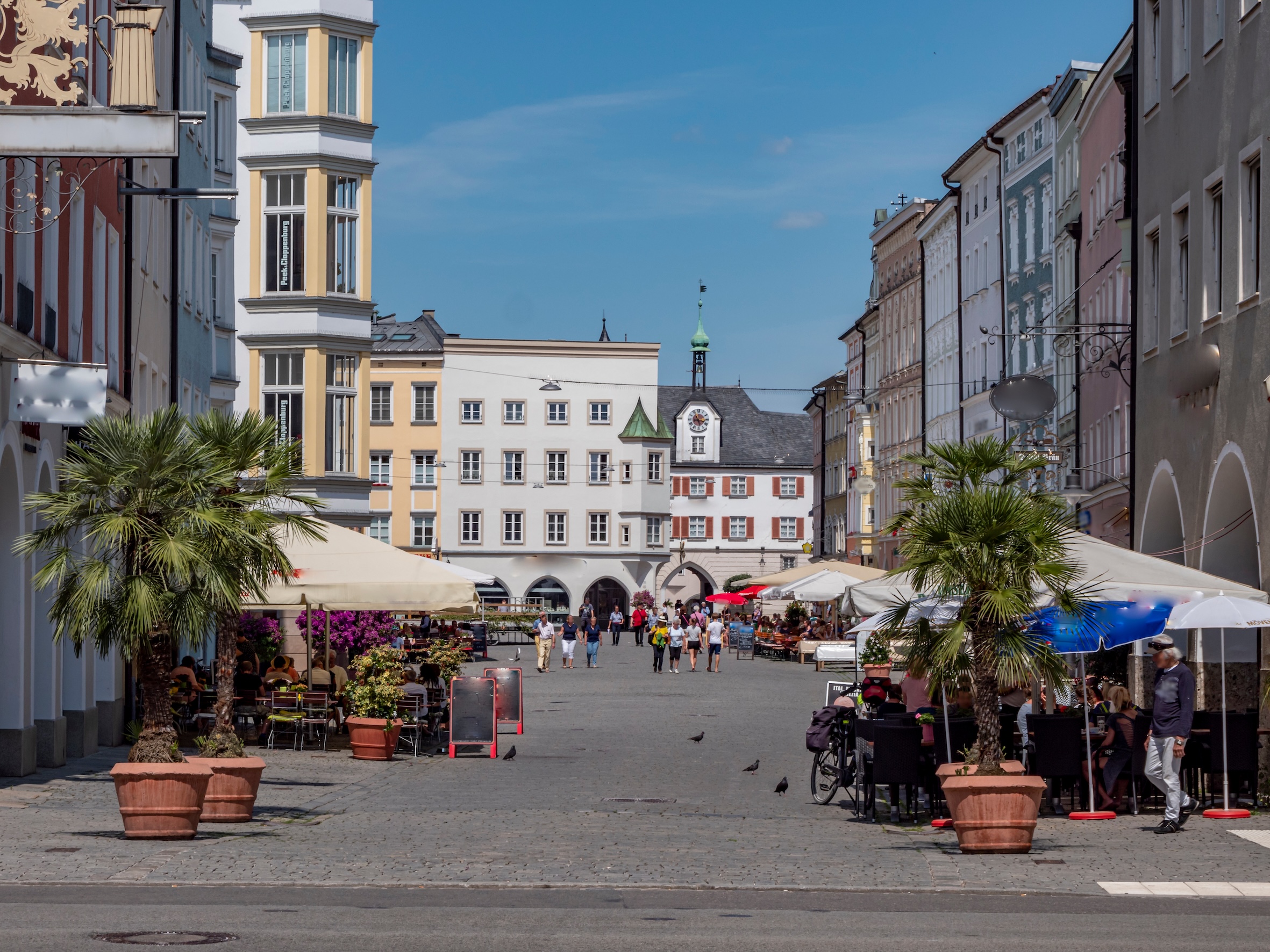Belebter Platz mit Straßencafés, Palmen in Kübeln und historischen Gebäuden im Hintergrund.