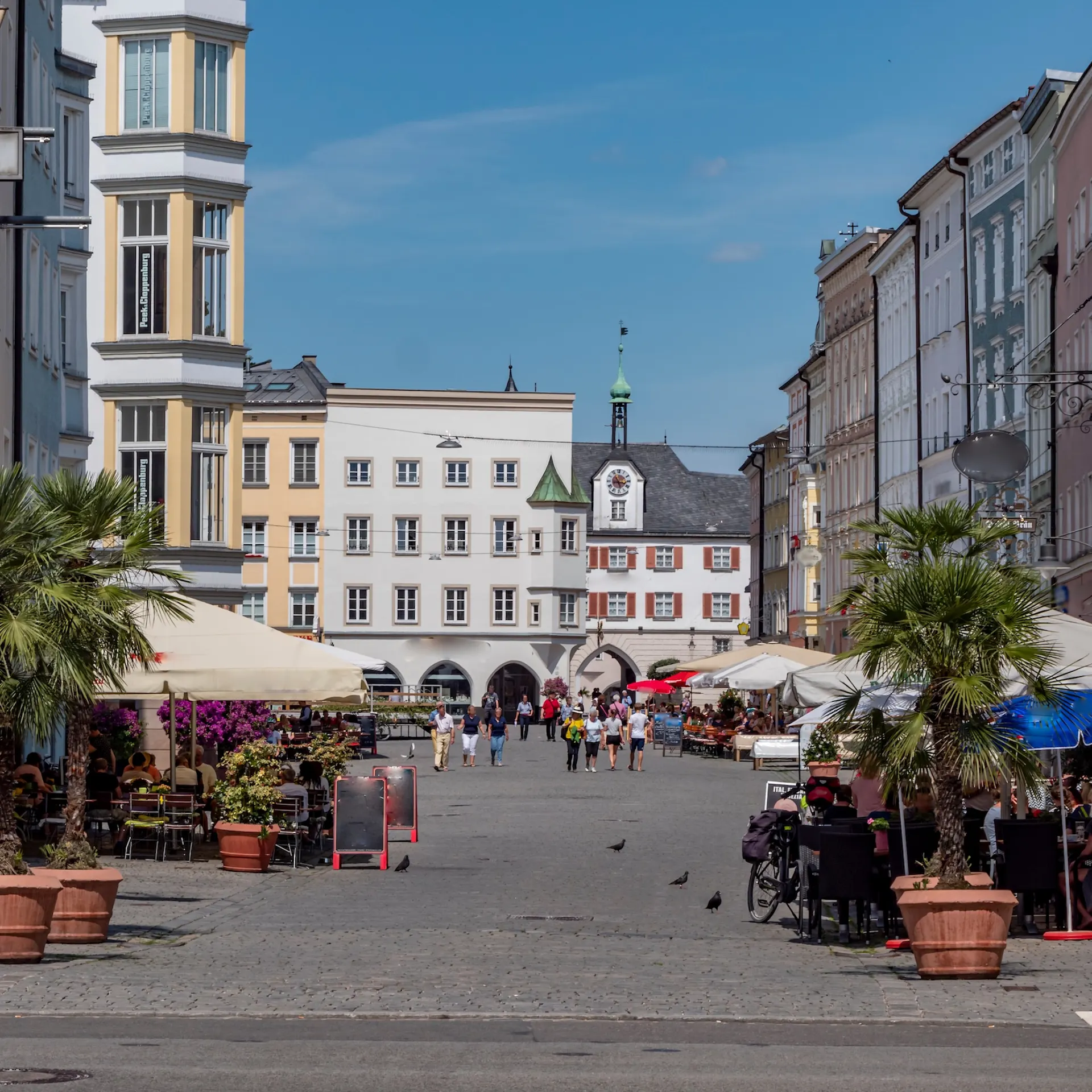 Belebter Platz mit Straßencafés, Palmen in Kübeln und historischen Gebäuden im Hintergrund.