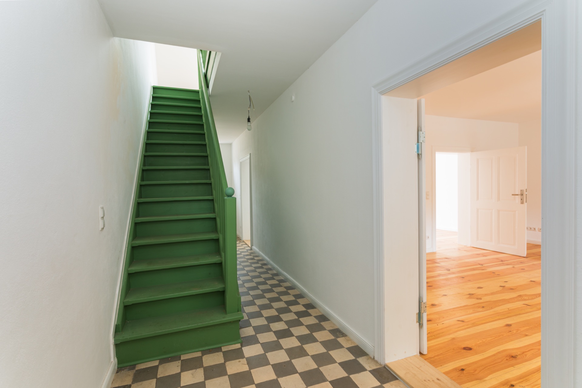 Flur mit grün gestrichener Treppe und kariertem Boden, daneben eine Tür zu einem hellen Raum.