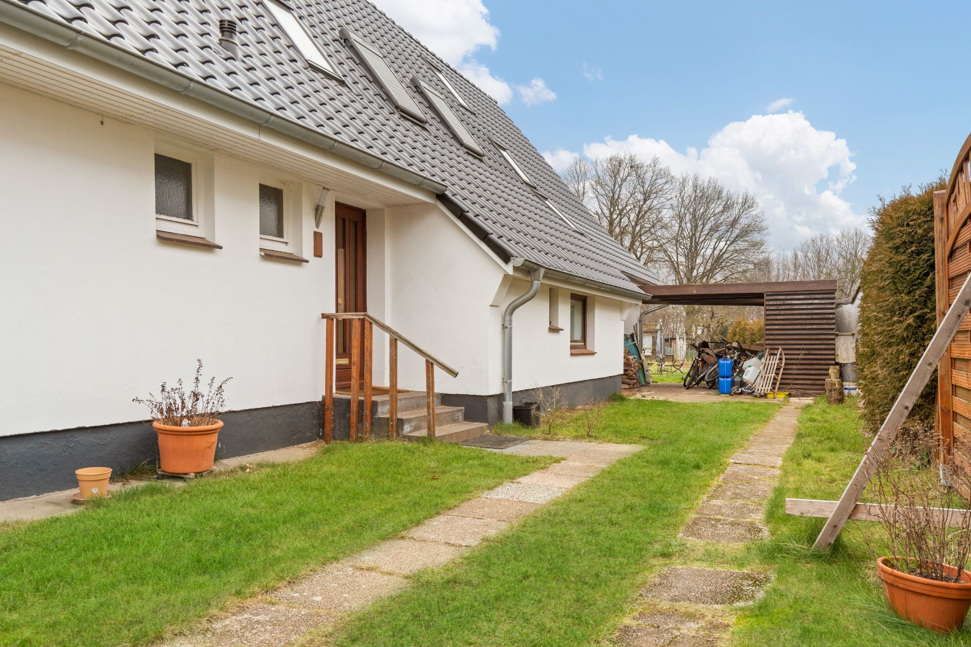 Weiße Hausfassade mit grauem Dach, Eingangstür, Rasen und überdachtem Abstellplatz im Garten.