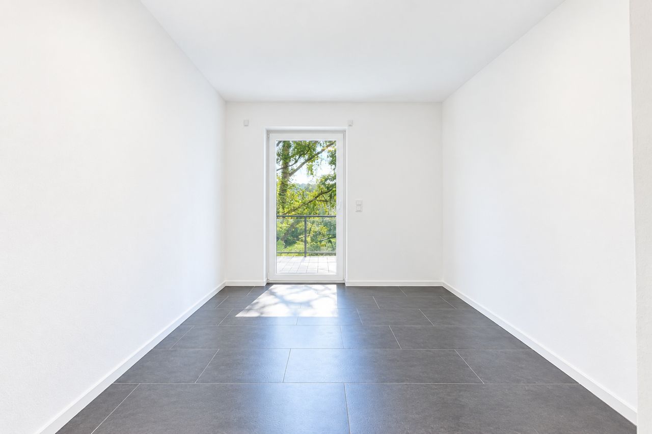 Heller leerer Raum mit großen dunklen Fliesen und Glastür zu einem Balkon oder Garten.