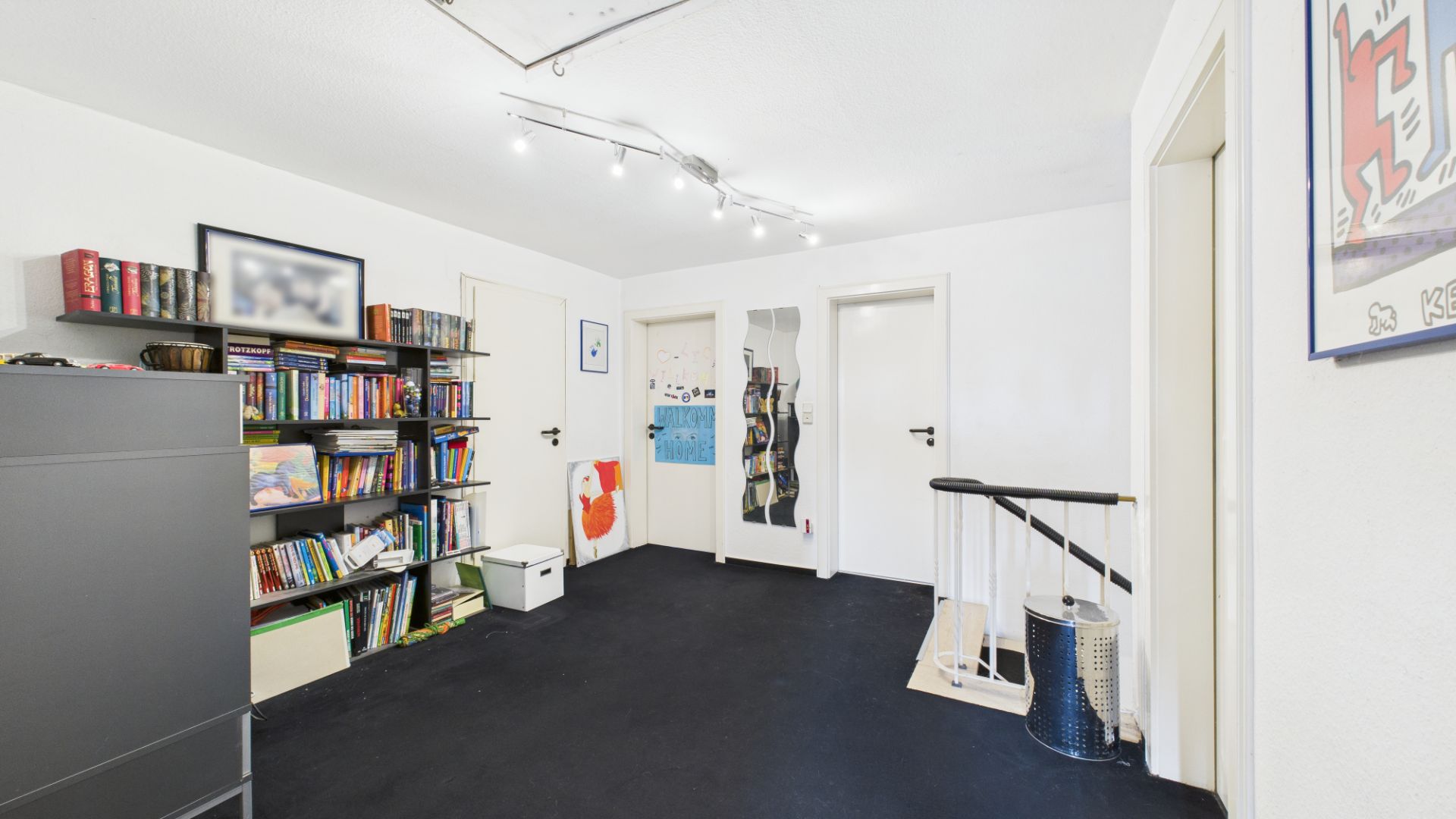 Heller Flur mit schwarzem Teppich, Bücherregal, drei Türen und Treppengeländer.