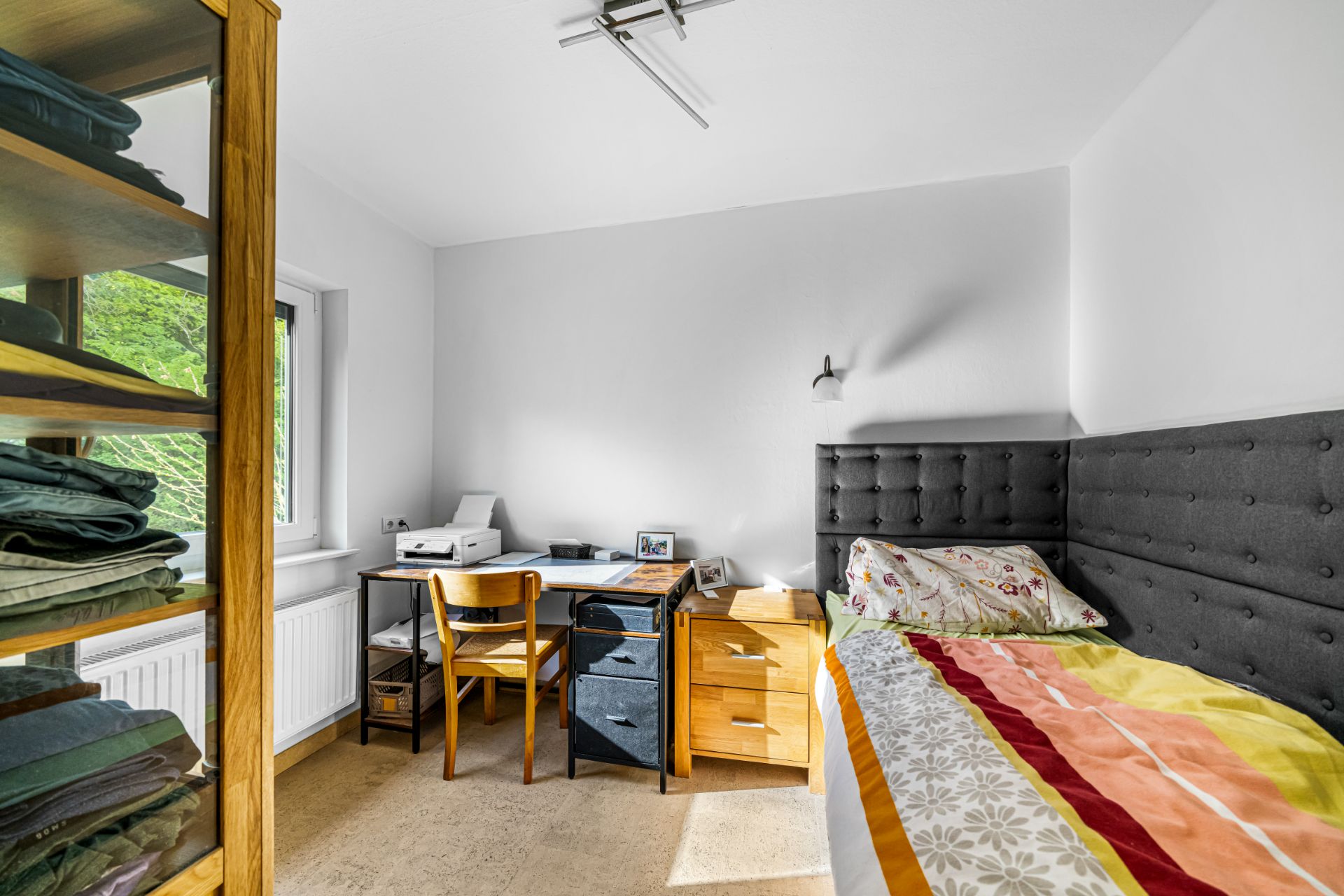 Kleines Schlafzimmer mit Einzelbett, Schreibtisch, Stuhl und Regal mit gefalteter Kleidung.