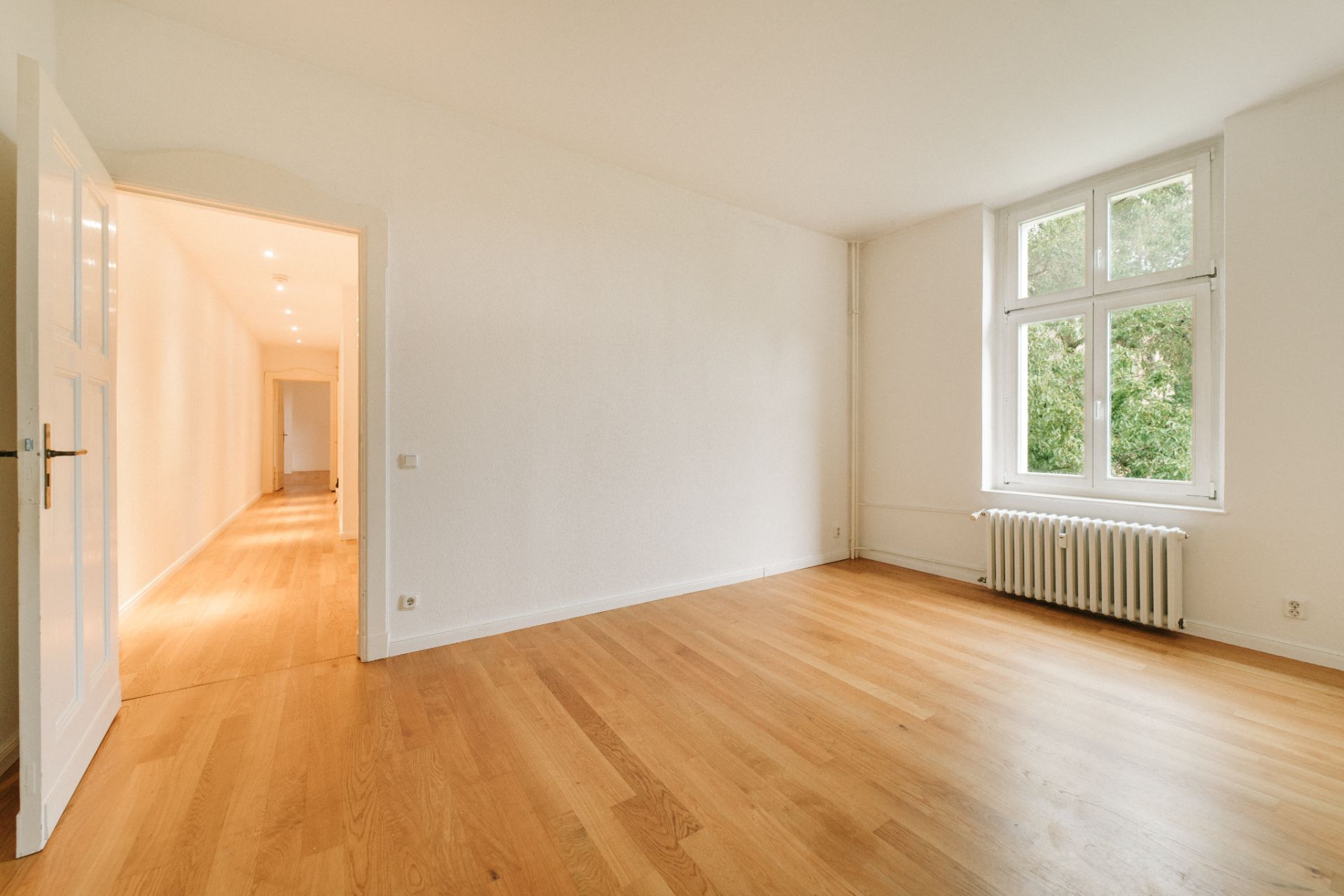 Leerer Raum mit Holzfußboden, großem Fenster und Heizkörper, Blick in einen langen Flur.