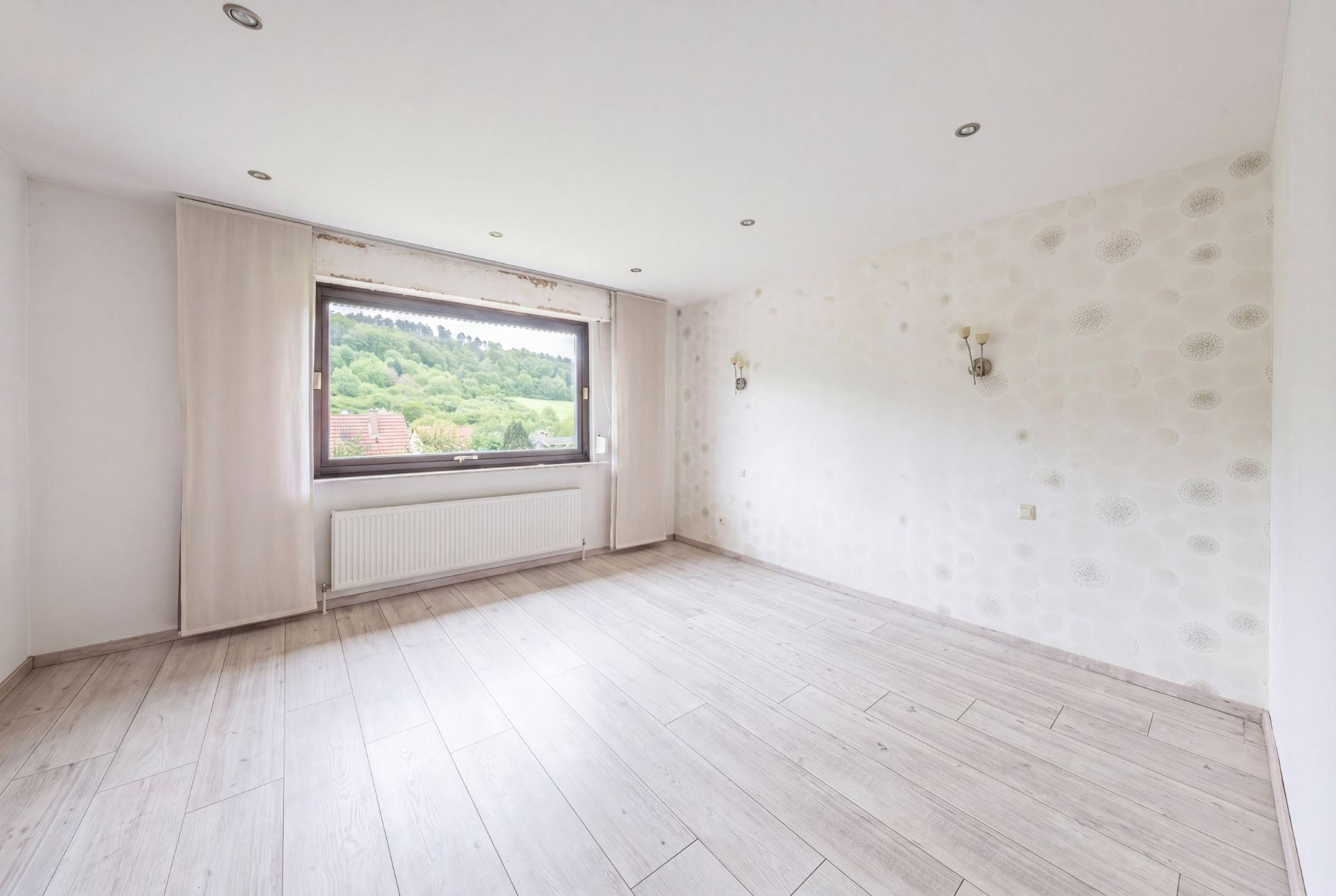 Helles, leeres Zimmer mit großem Fenster, Holzfußboden und Blick auf grüne Landschaft.