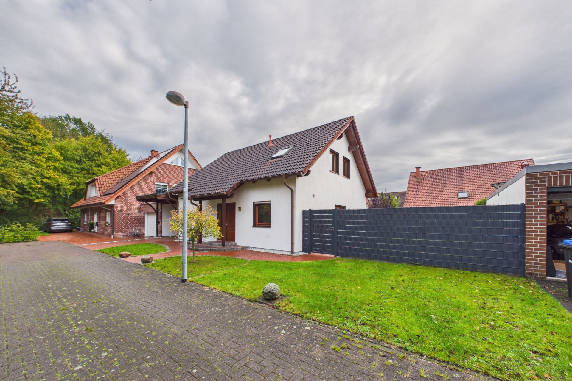 Modernes Einfamilienhaus mit Garten, grauem Zaun und gepflasterter Einfahrt bei bewölktem Himmel.