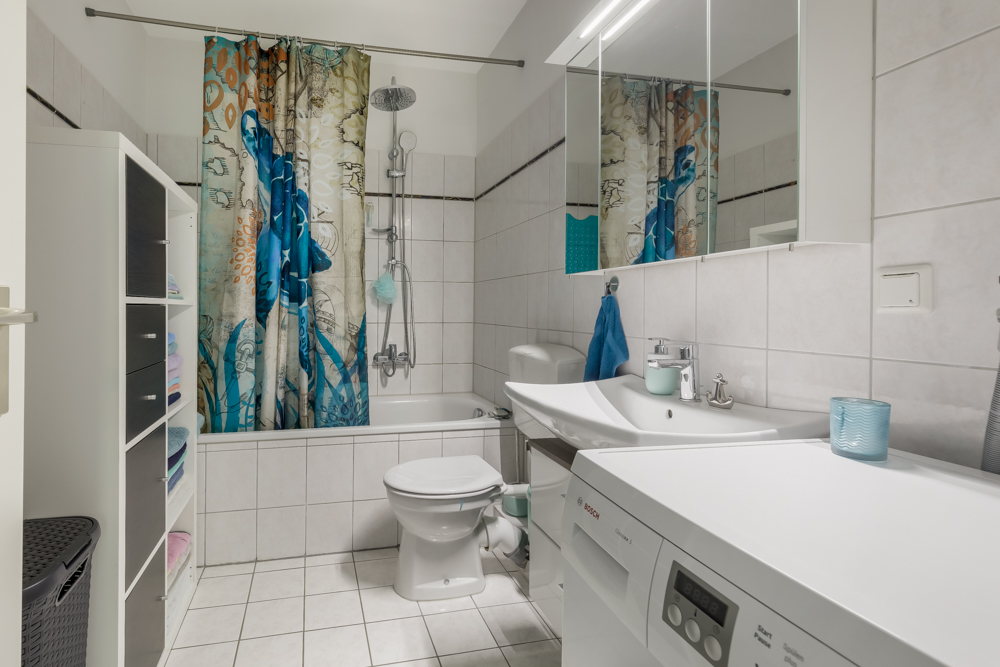 Modernes Badezimmer mit Waschmaschine, Badewanne, Duschvorhang, WC und Regal mit Handtüchern.