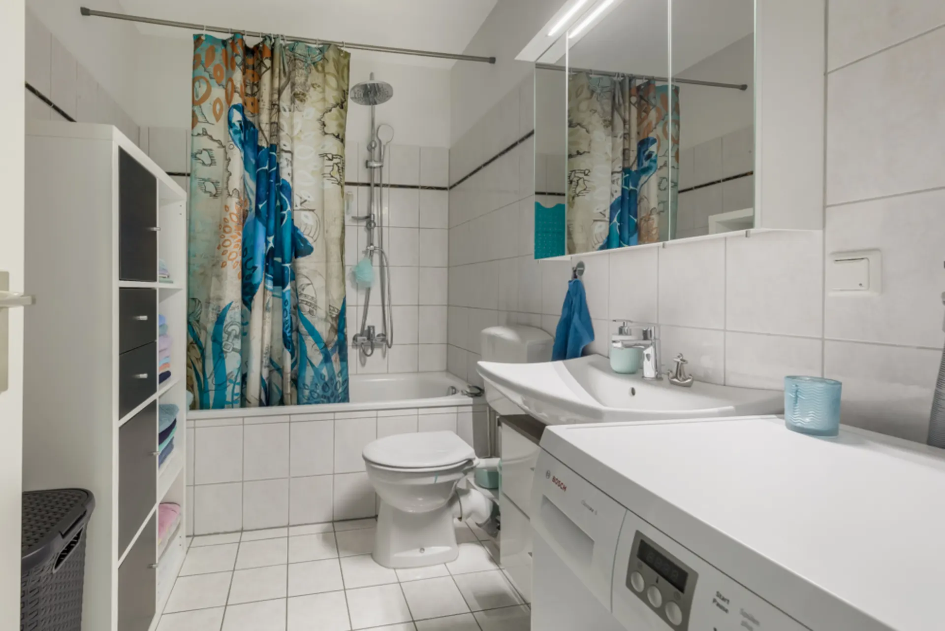 Modernes Badezimmer mit Waschmaschine, Badewanne, Duschvorhang, WC und Regal mit Handtüchern.