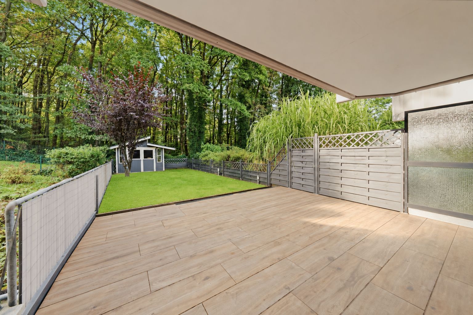 Überdachte Terrasse mit Holzboden, angrenzend an einen Garten mit kleinem Gartenhaus und Zaun.