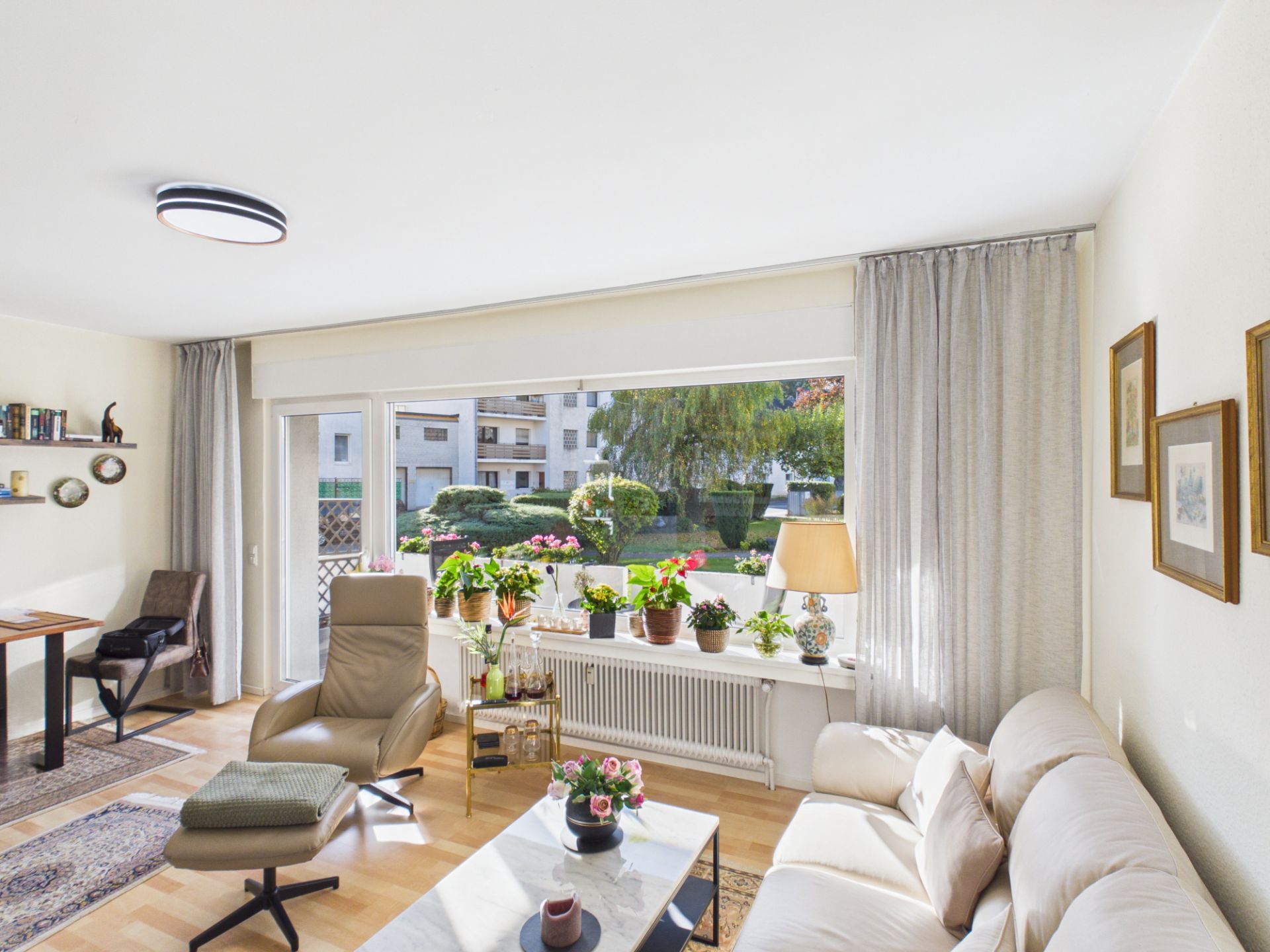 Helles Wohnzimmer mit Sofa, Sessel, Couchtisch und vielen Pflanzen vor einem großen Fenster.
