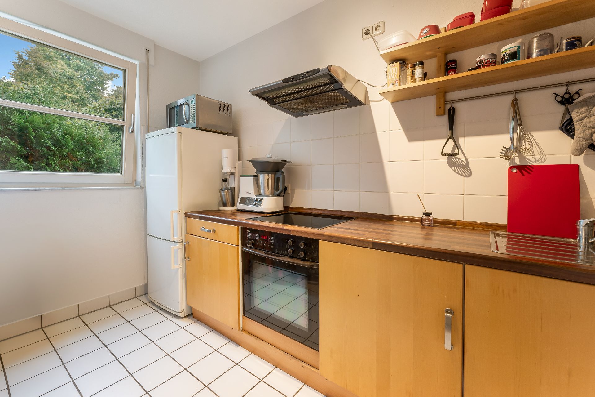 Helle Küche mit Holzschränken, Herd, Kühlschrank und Küchenutensilien, Fenster mit Blick ins Grüne.