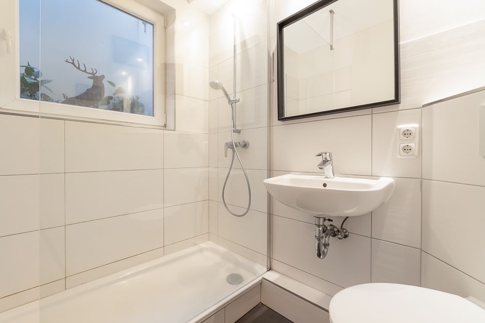 Modernes, hell gefliestes Bad mit Dusche, Waschbecken, Spiegel und Fenster.