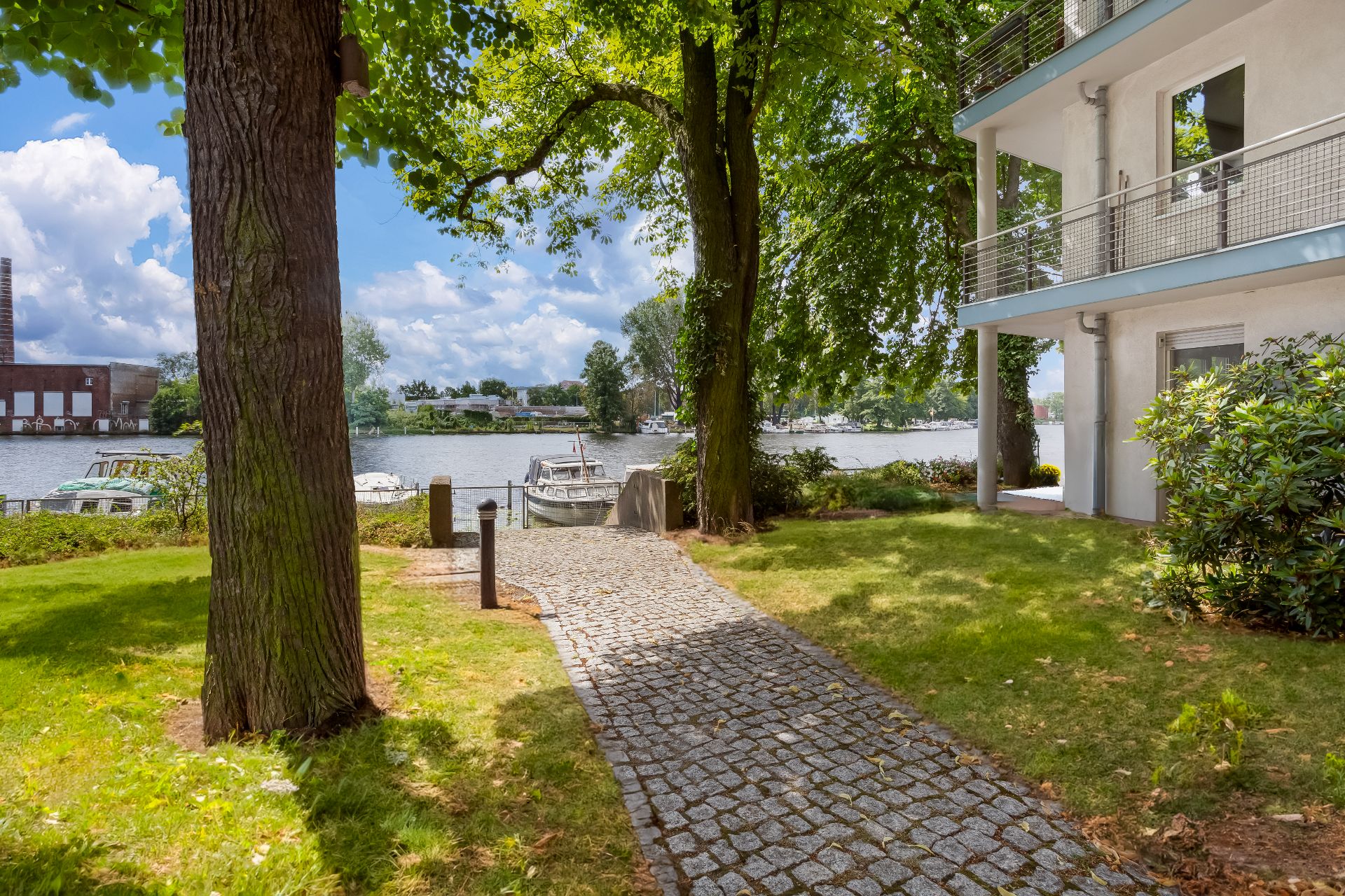 Gepflasterter Weg führt durch Garten mit Bäumen zu einem Fluss, rechts ein Wohngebäude mit Balkon.