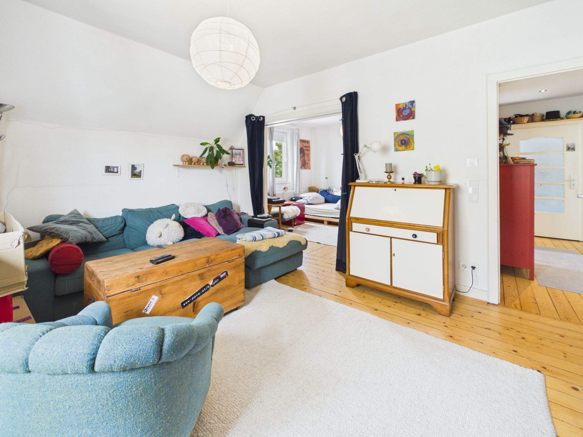 Helles Wohnzimmer mit blauem Sofa, Holztruhe, Teppich und Zugang zu weiteren Räumen.
