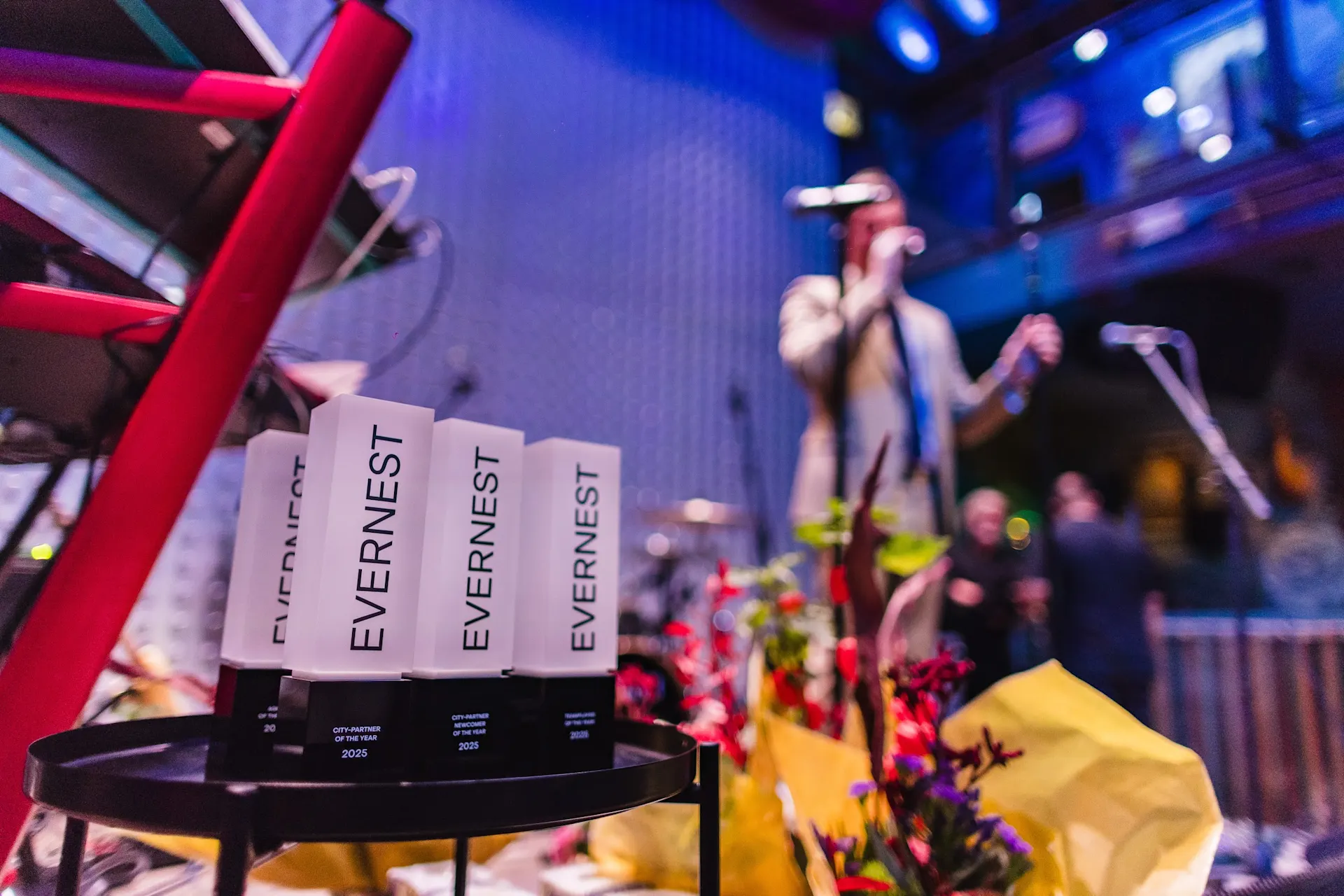 Ein Tisch mit Evernest-Trophäen und Blumen, im Hintergrund eine Person an einem Mikrofon.