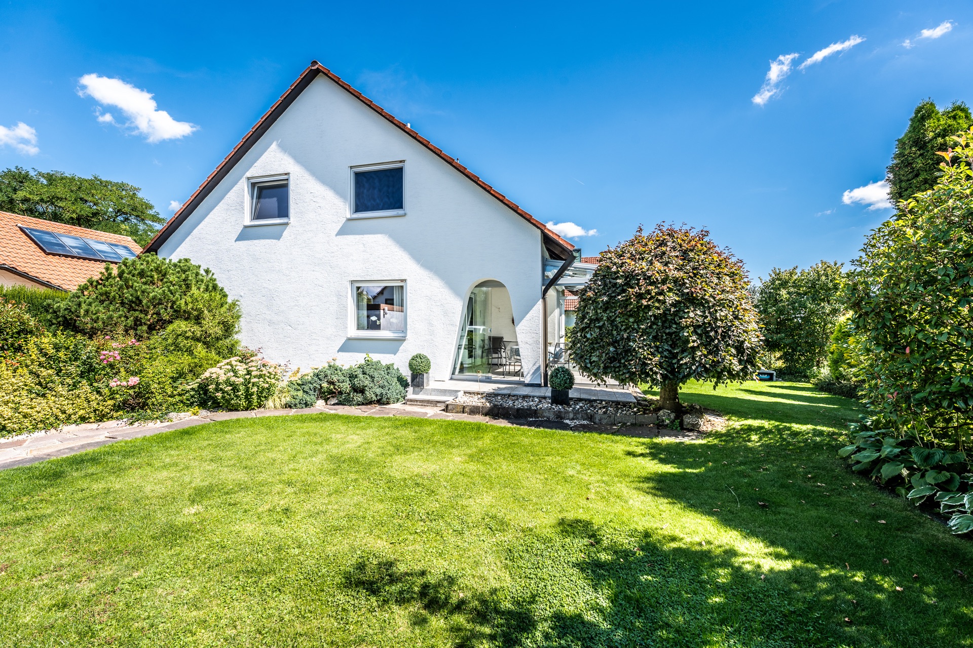 Weißes Einfamilienhaus mit Garten, Terrasse und Bäumen an einem sonnigen Tag.