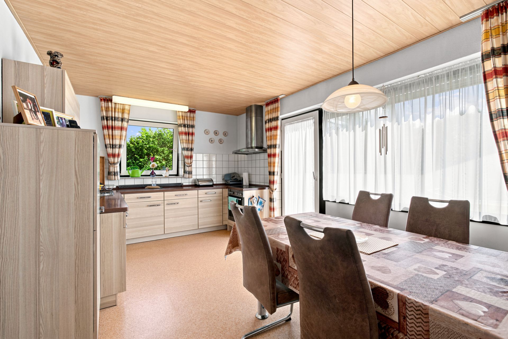 Helle Küche mit Esstisch, vier Stühlen, großen Fenstern und Gardinen.